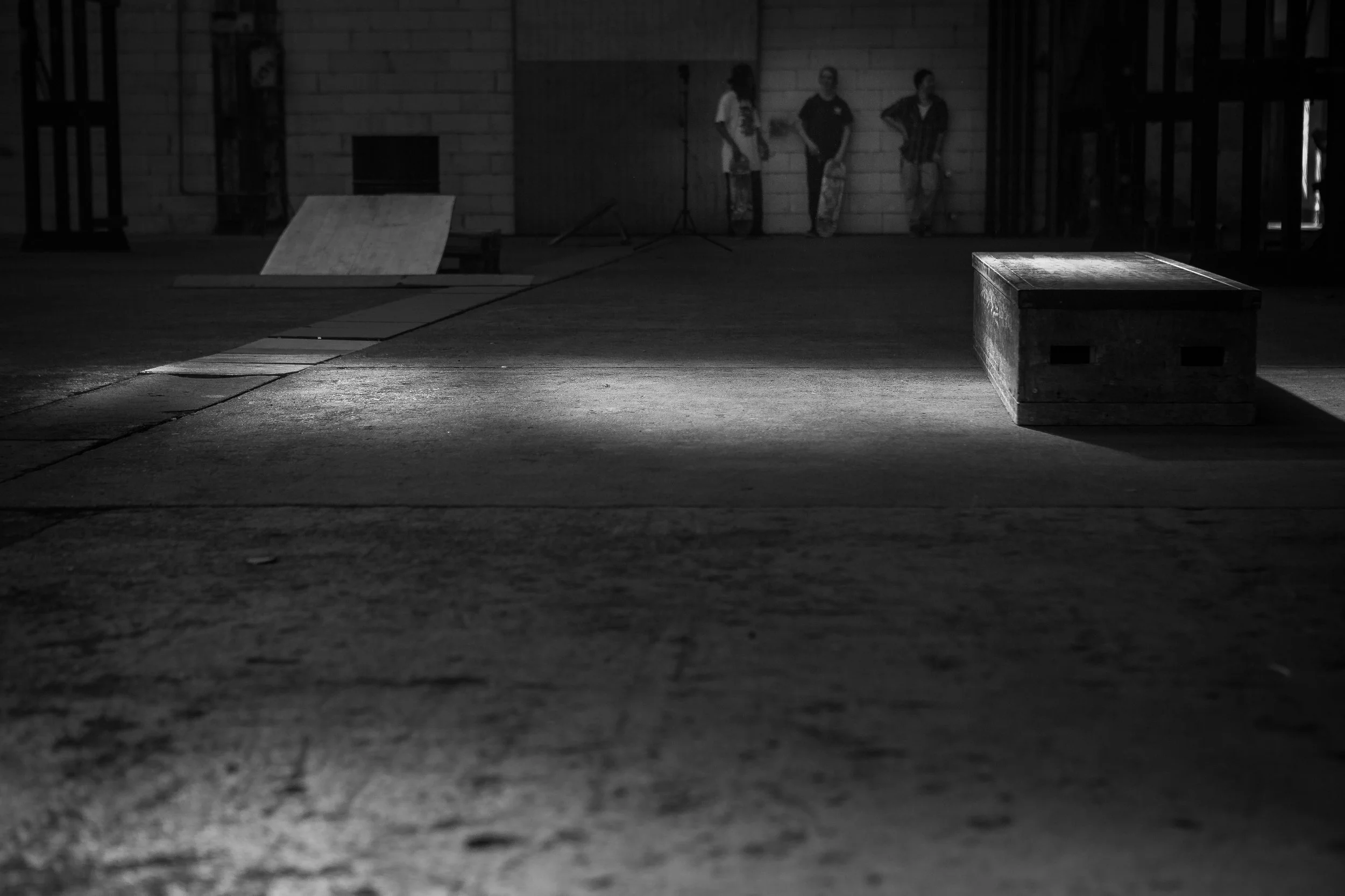 Dunkler Innenraum mit drei stehenden Personen im Hintergrund, eine auf einem Skateboard, und einem Holz-Box-Element im Vordergrund, Licht- und Schatteneffekte auf dem Boden.