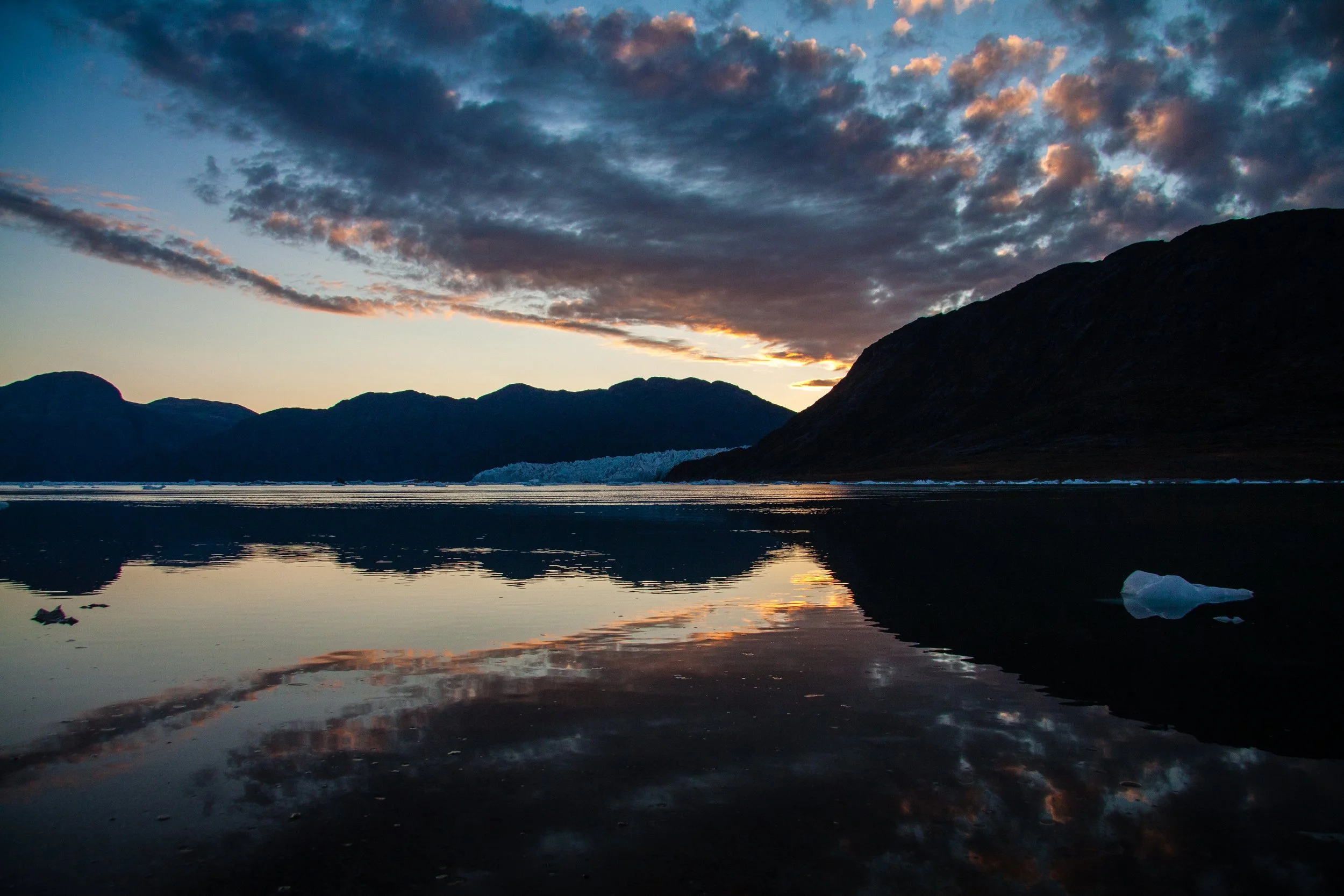 Sonnenuntergang über Bergen und Wasser, Reflexion der Wolken im Wasser, kleines Stück Eis im Vordergrund.