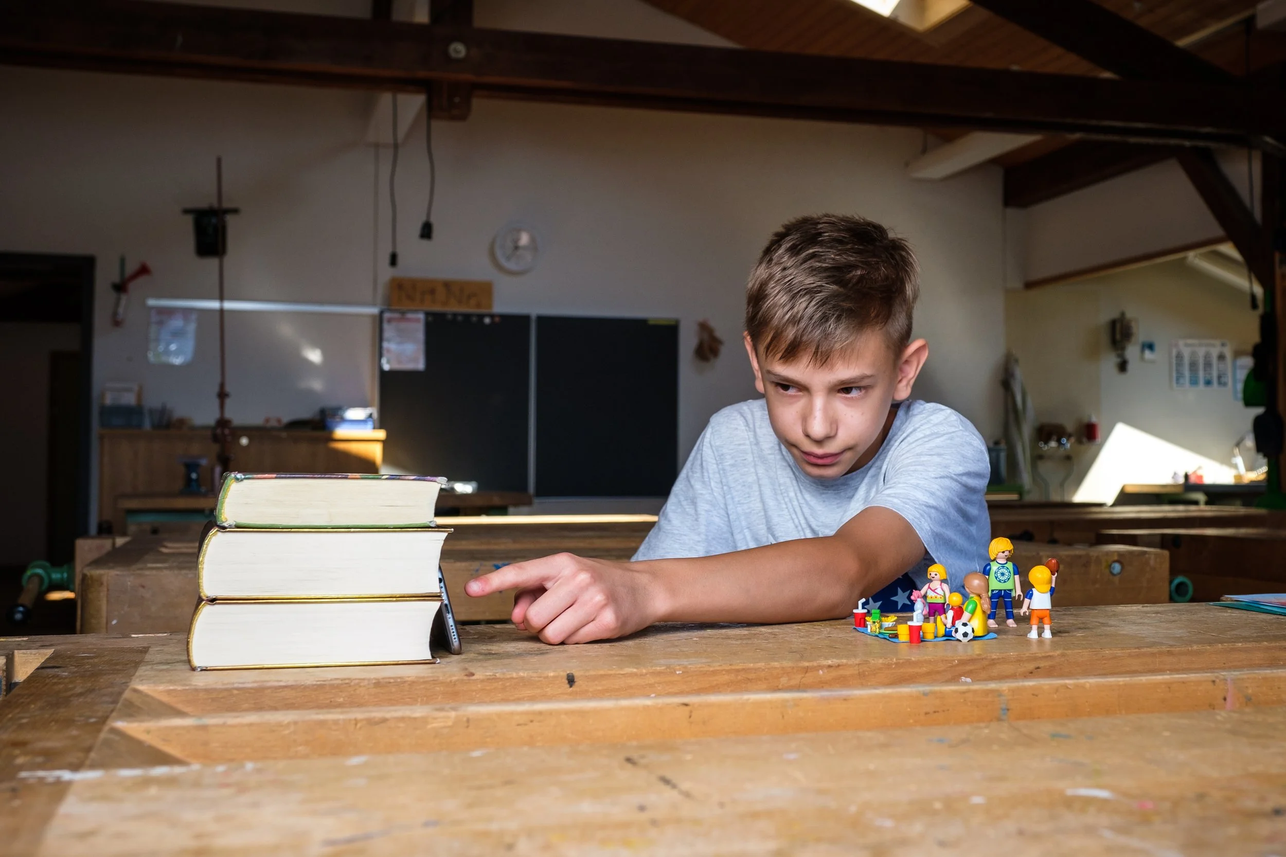 Ein Junge liegt an einem Holztisch, schaut auf Lego-Figuren und ein Buch im Vordergrund, in einem Holzraum mit dunklem Dach.