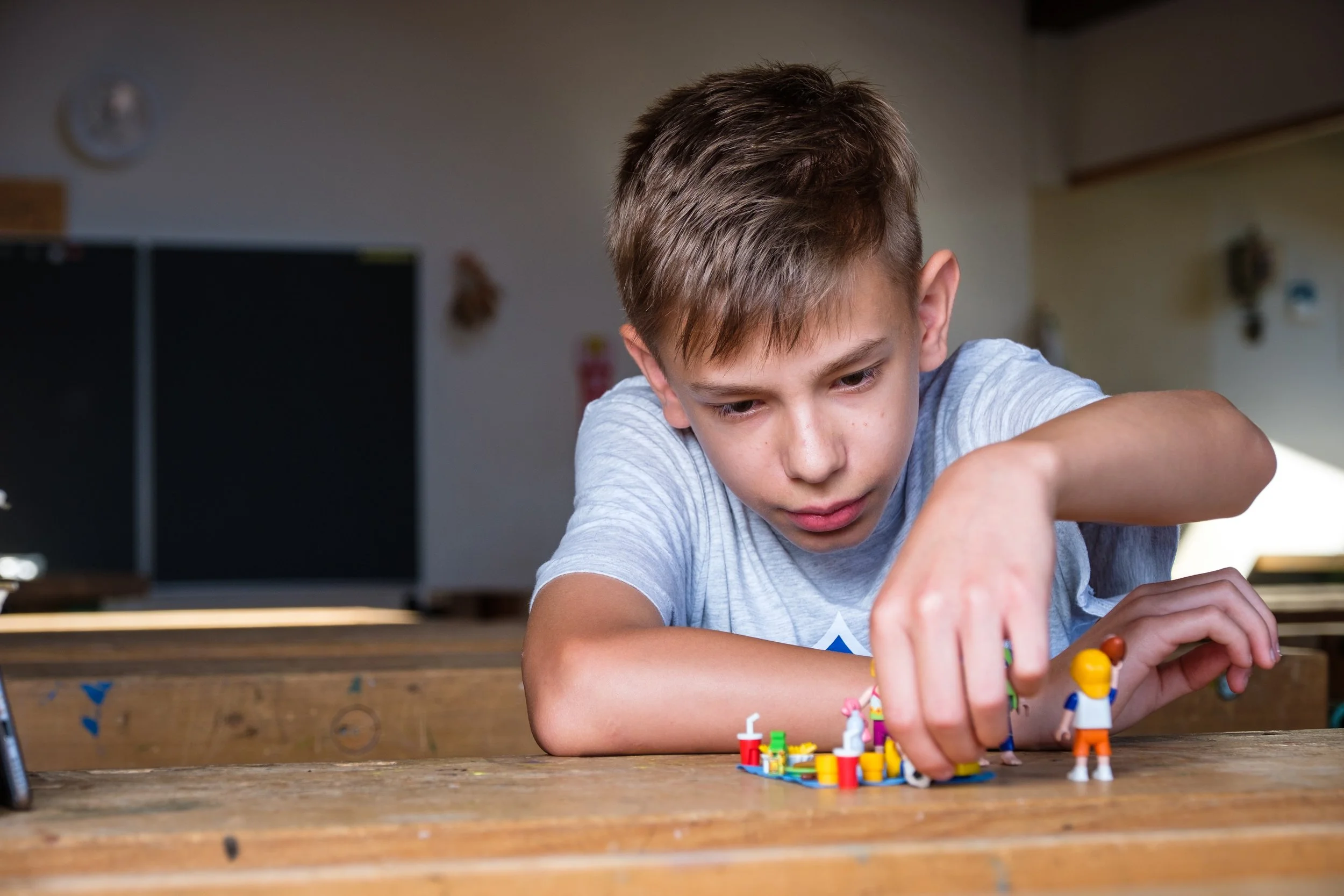 Junge spielt mit Lego-Figuren auf einem Holztisch in einem Klassenraum.