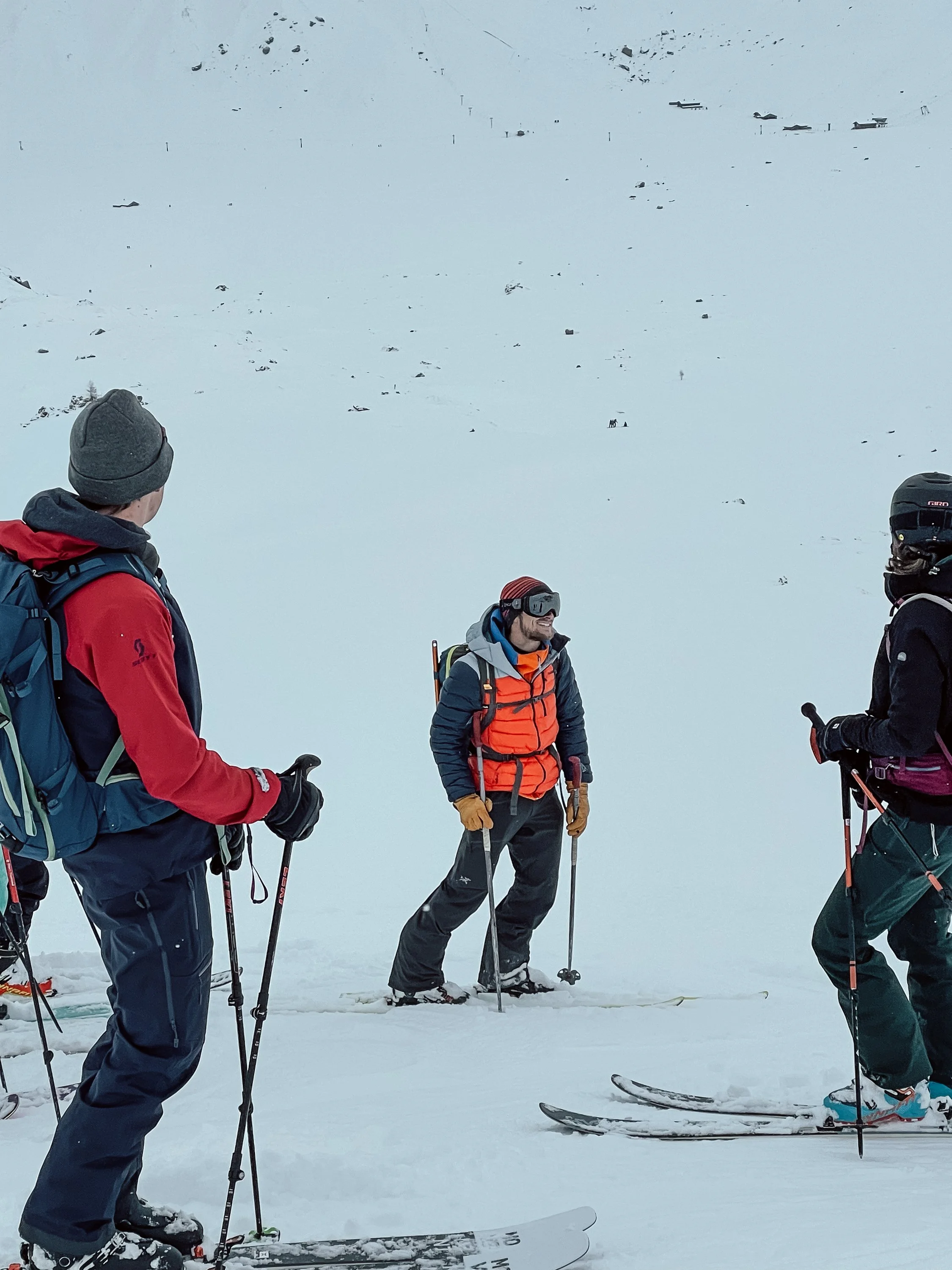 Drei Skifahrer in Winterkleidung auf verschneitem Berg, einer spricht mit den anderen, im Hintergrund Schnee und Skihütten