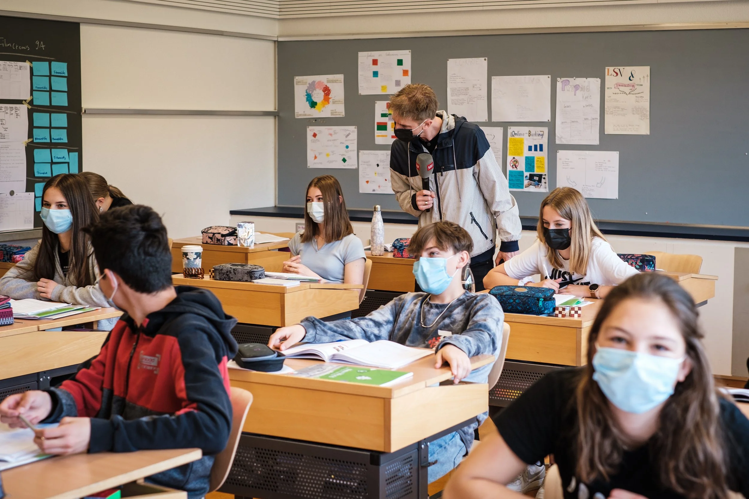 Schüler in einem Klassenzimmer mit Masken, ein Lehrer mit einem Mikrofon steht während des Unterrichts.