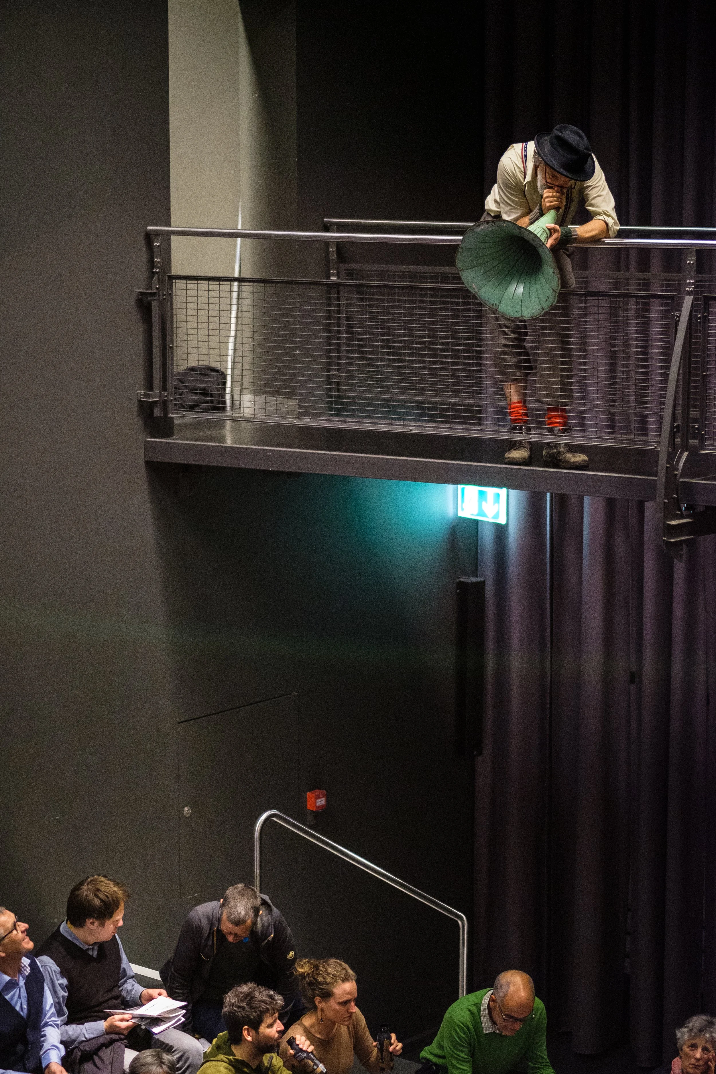 Ein Mann trägt einen Hut und einen Anzug, steht auf einem Balkon, lehnt sich vor und spricht in eine alte Grammophon-Schallbox.