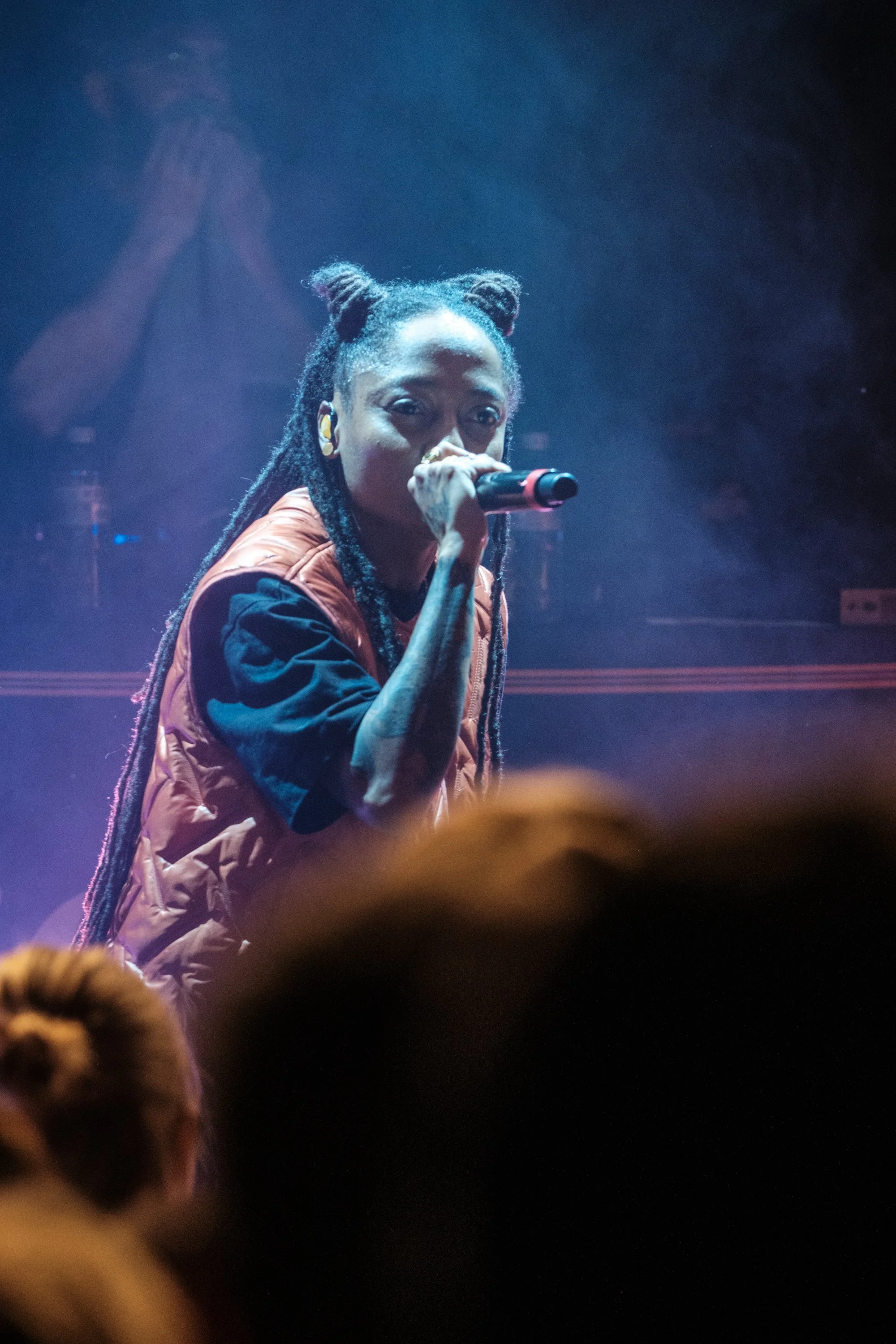 Eine junge Frau mit Dreadlocks singt in einem Konzert mit einem Mikrofon, im Hintergrund erkennbare Person mit verschränkten Händen.