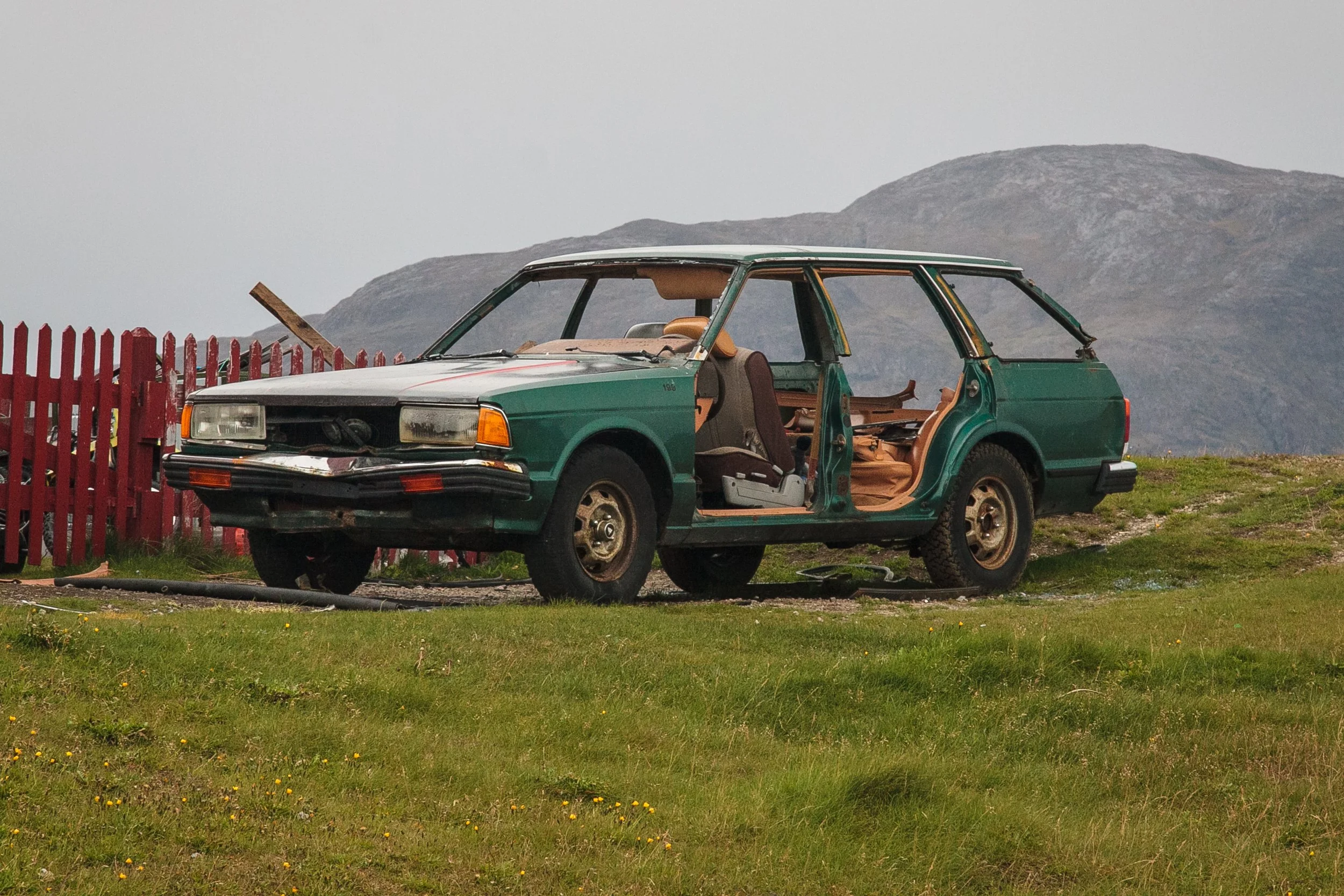 Ein verlassener, beschädigter grüner Kombi auf einer grünen Wiese mit Bergen im Hintergrund.