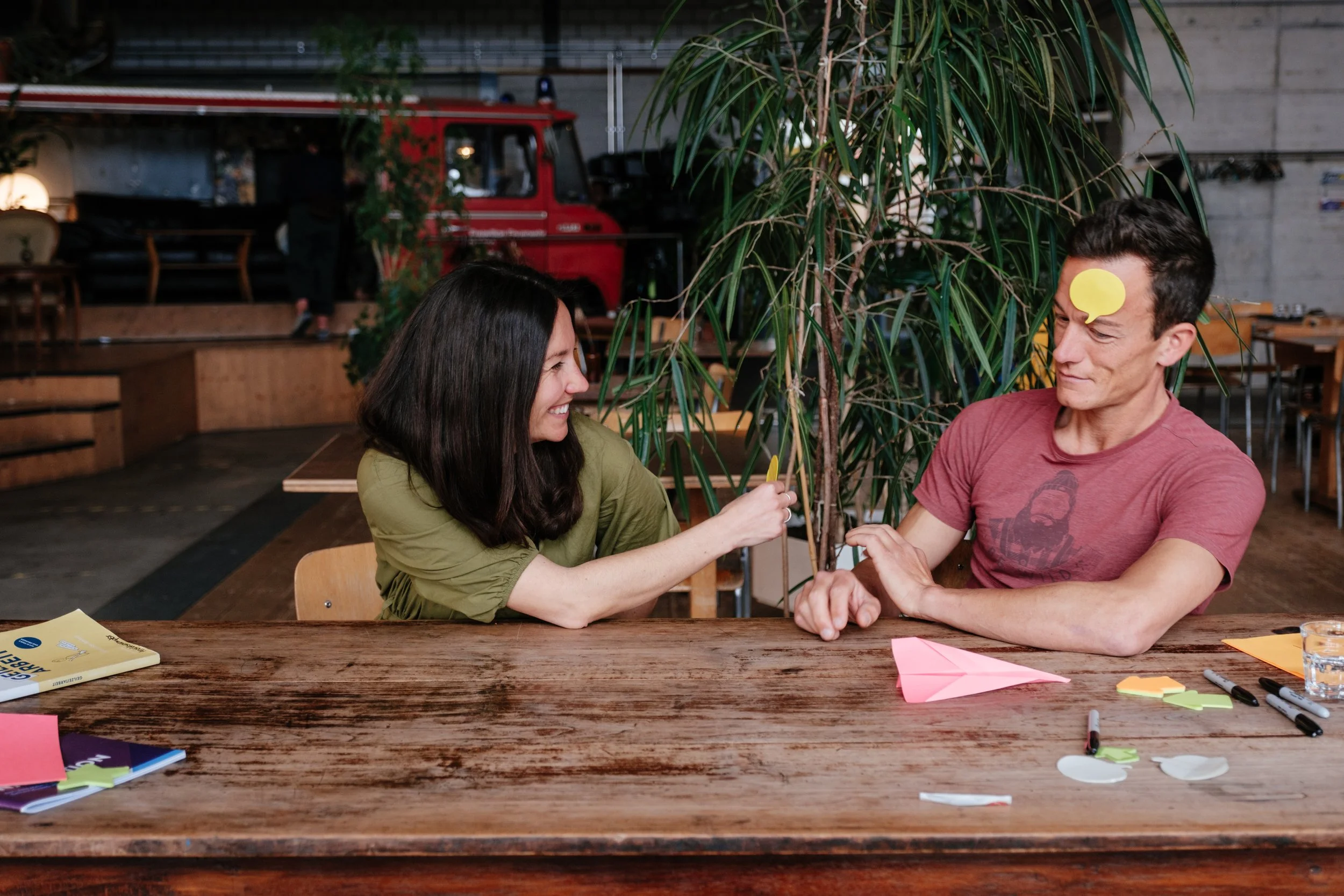 Zwei Menschen sitzen an einem Holztisch, lachen und spielen mit Papierflugzeugen in einem Café oder Restaurant, im Hintergrund Pflanzen und eine Wand mit einem roten Feuerwehrauto.