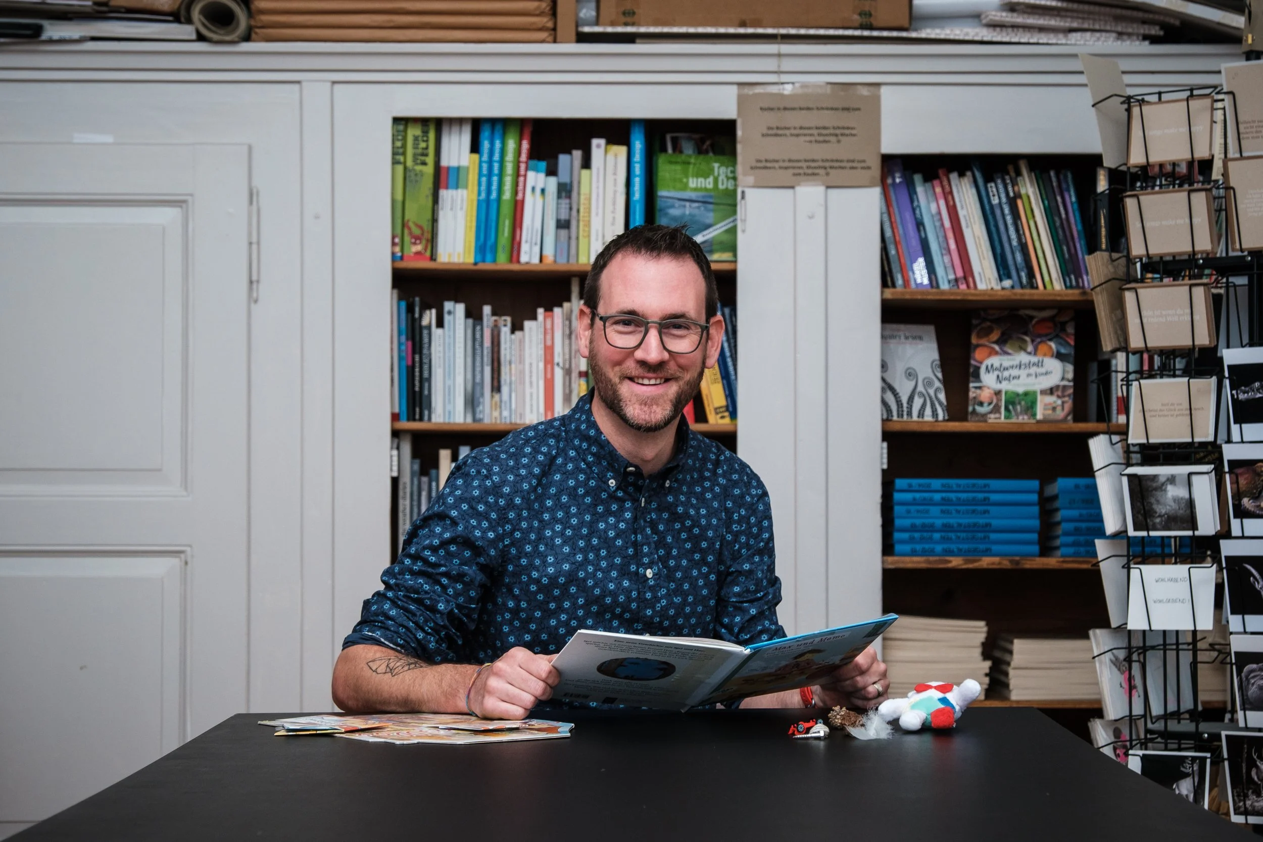Ein lachender Mann mit Brille sitzt an einem schwarzen Tisch in einer Buchhandlung oder Bibliothek, liest ein Magazin und umgeben von Büchern, Postkarten und kleinen Taschen. Im Hintergrund sind Bücherregale mit bunten Büchern und Regalschildern.