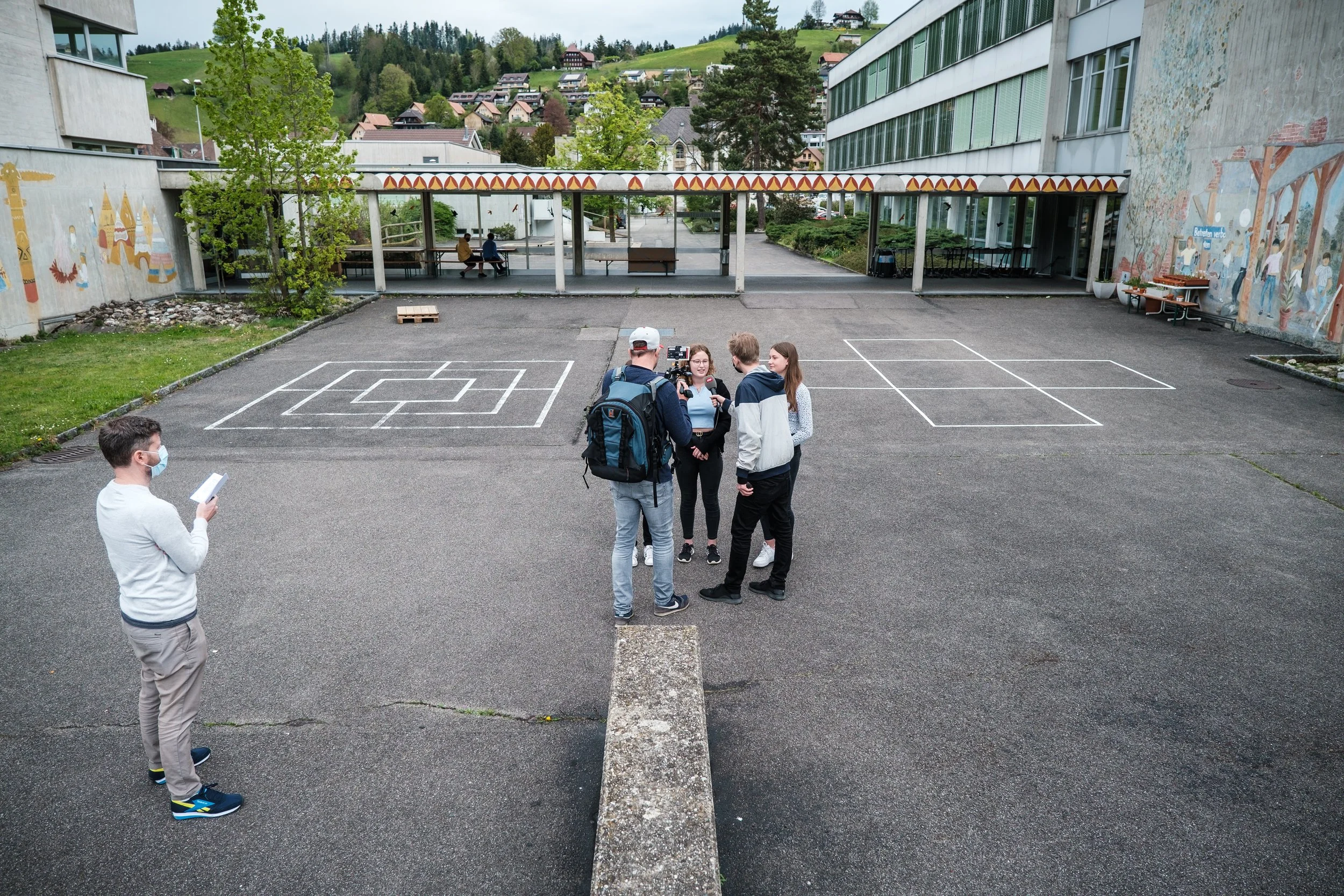 Gruppen von jungen Menschen im Gespräch auf einem Schulhof, einige tragen Masken, im Hintergrund bunte Wandkunst, Schulgebäude und eine grüne, hügelige Landschaft. Ein Mann steht links mit einem Notizblock, die Gruppe scheint ein Interview zu geben.