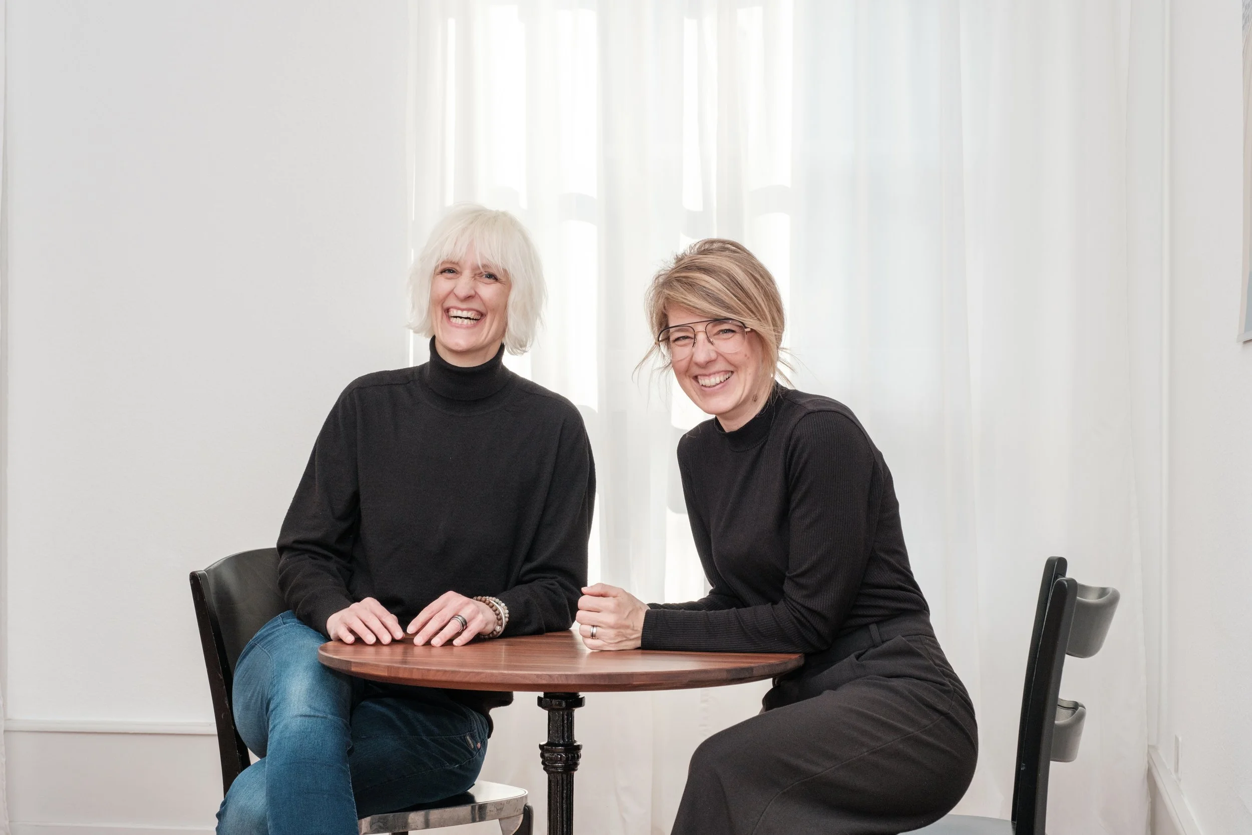 Zwei lachende Frauen sitzen an einem runden Holztisch vor weißen Vorhängen.