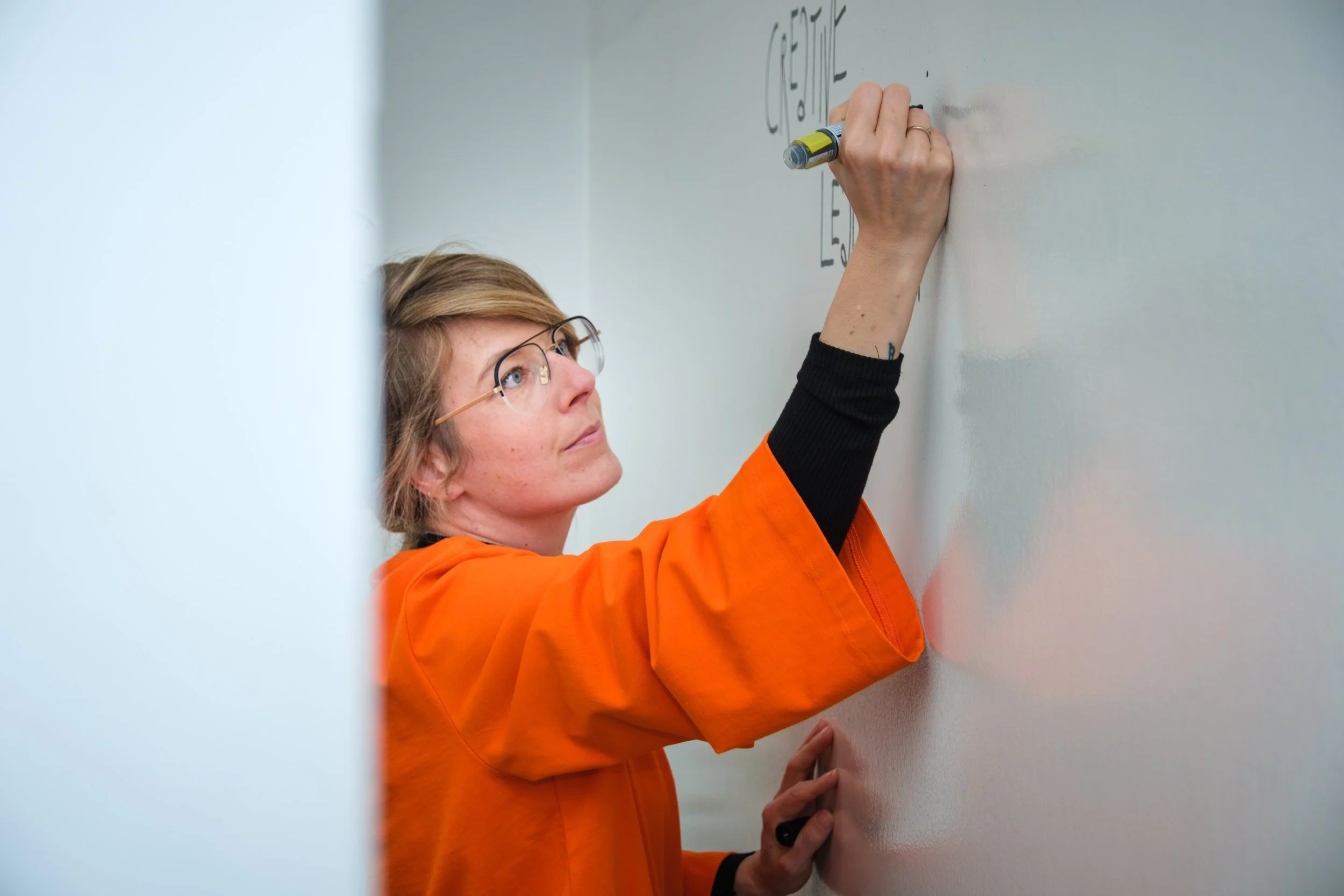 Frau schreibt auf eine weiße Tafel mit einem schwarzen Marker, trägt eine orangejacke und Brille.