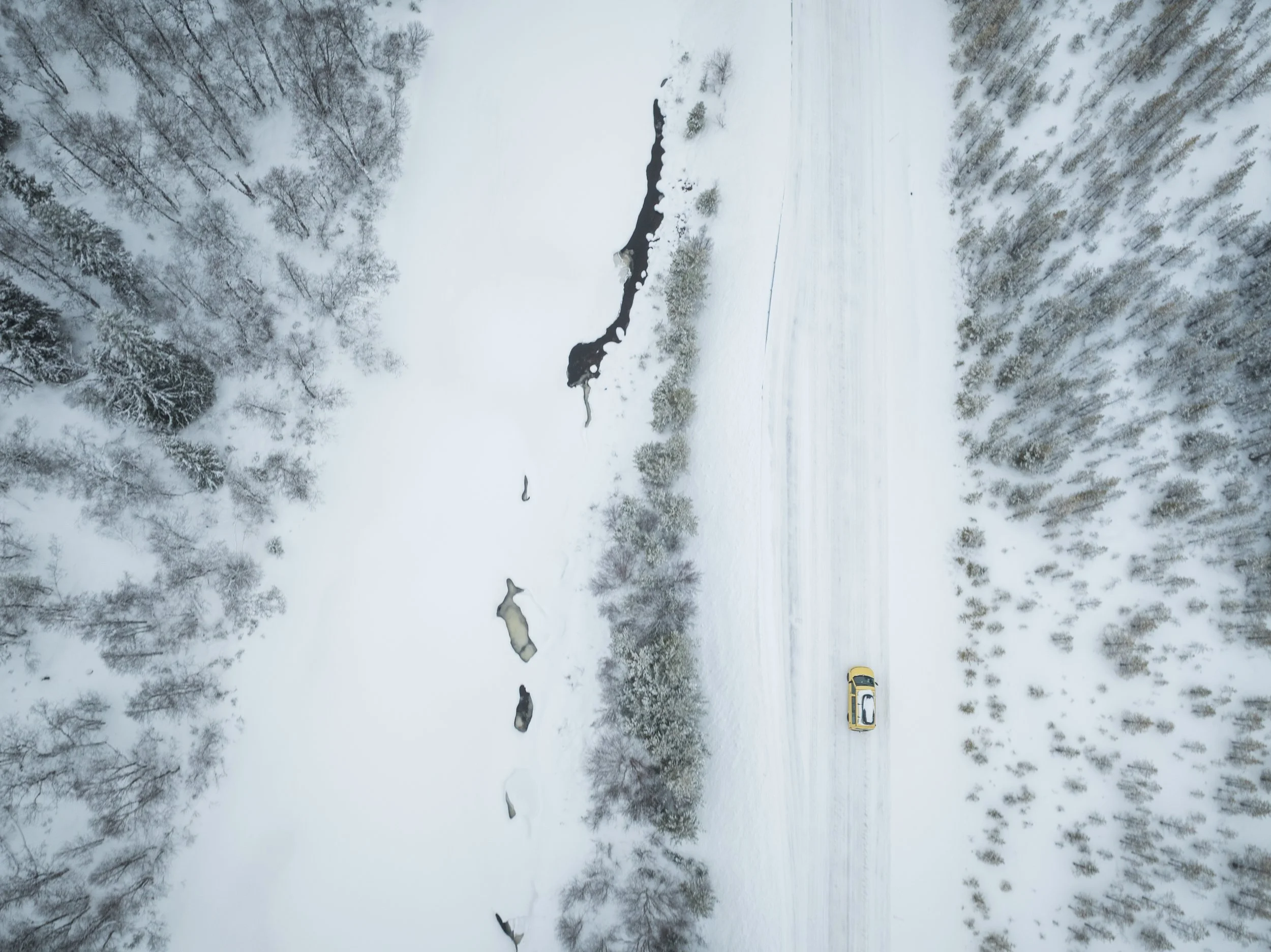 Vogelperspektive auf eine verschneite Winterlandschaft mit einem gelben Auto auf einer schneebedeckten Straße, umgeben von verschneiten Bäumen und gefrorenen Seen.