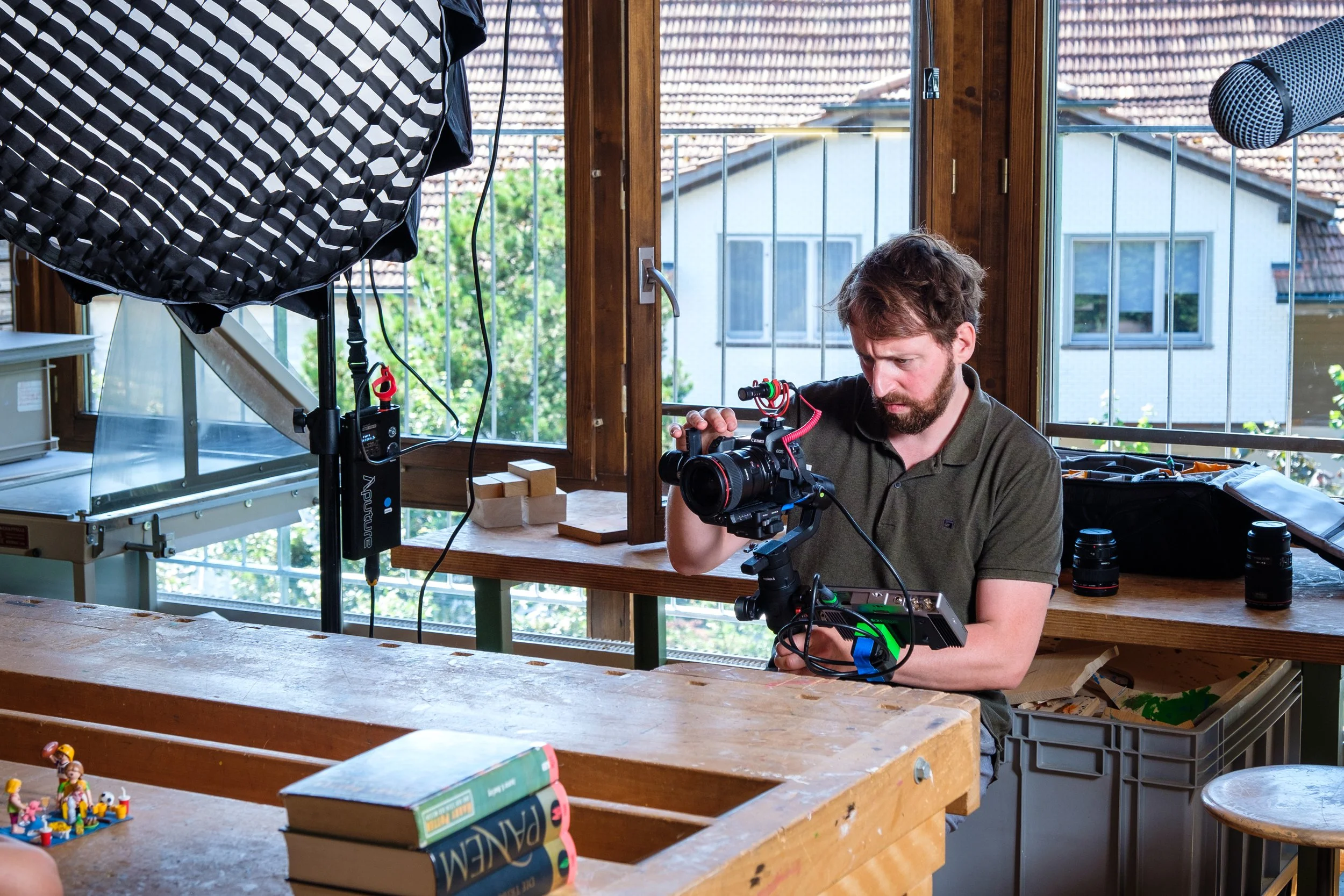 Mann richtet professionelle Kamera auf einem hell erleuchteten Raum mit großen Fenstern und Rattan-Möbeln. Es gibt Beleuchtungs- und Kameratechnik auf einem Holzschreibtisch, sowie Bücher und Spielzeug auf dem Tisch im Vordergrund.