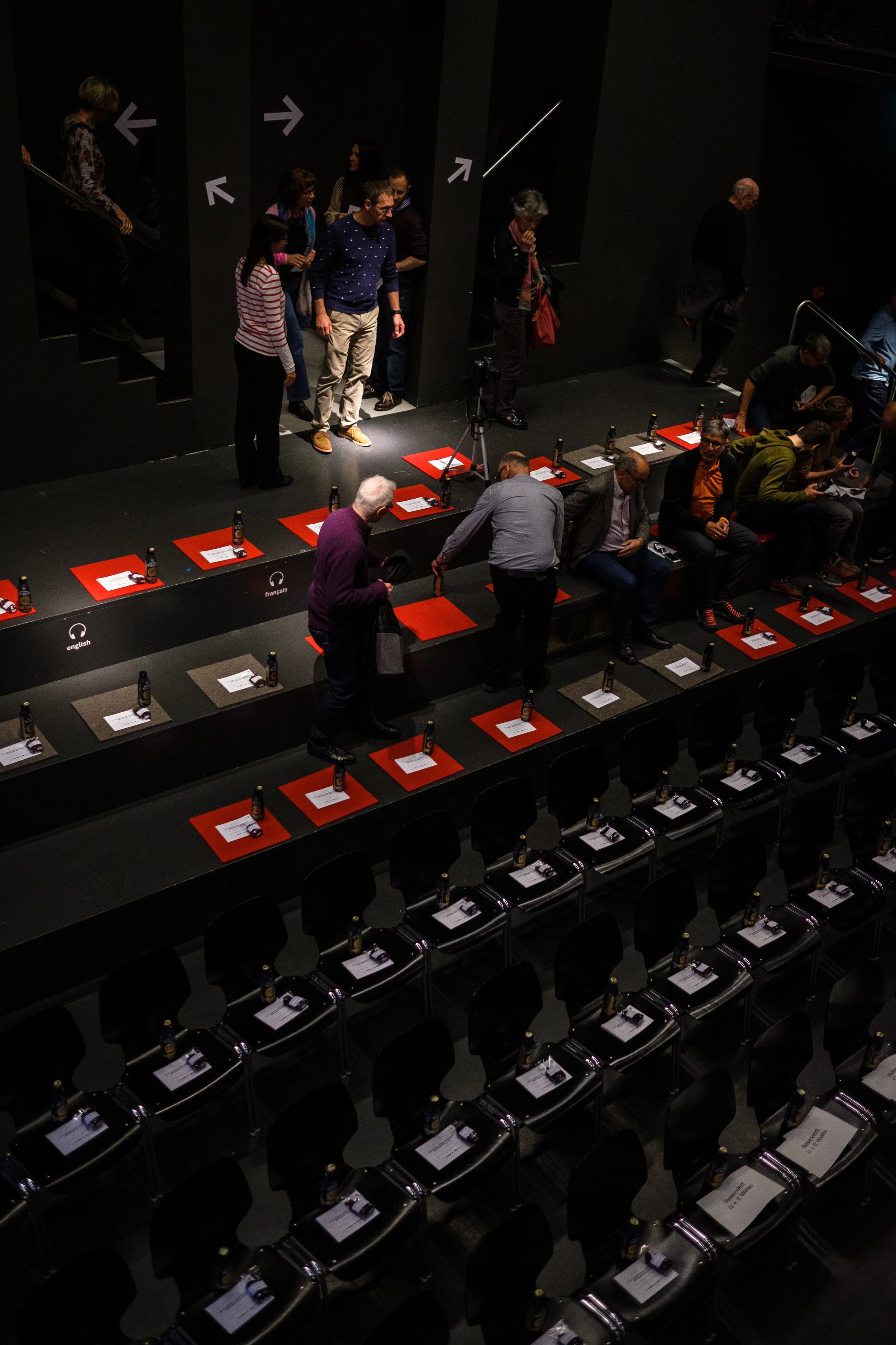 Menschen warten in einem dunklen Veranstaltungsraum, Sitzplätze mit roten Unterlagen sind vorbereitet, einige Personen sind bereits auf der Bühne, während andere noch im Raum stehen.