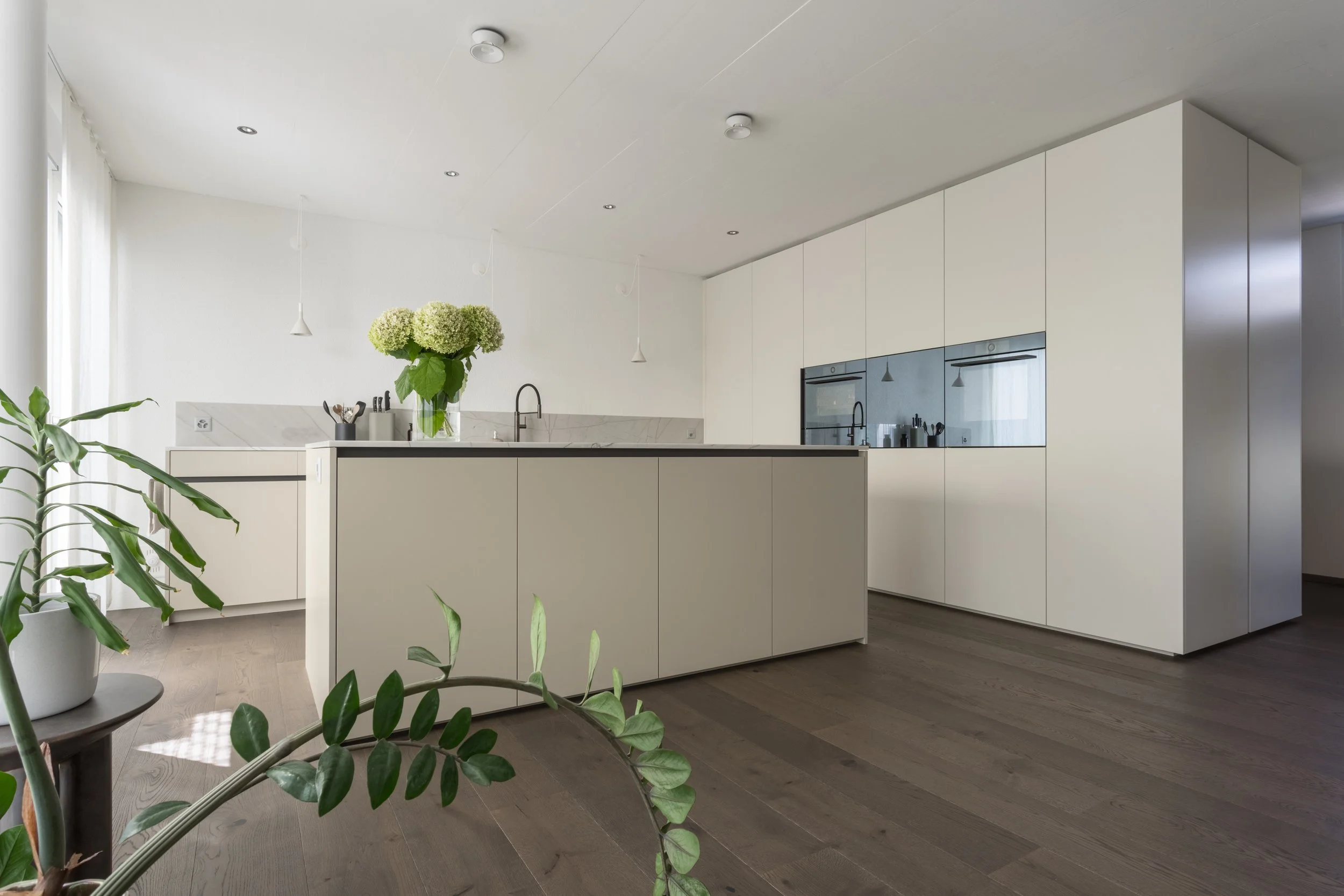 Moderne, minimalistische weiße Küche mit Kücheninsel, Pflanzen und Holzfußboden.