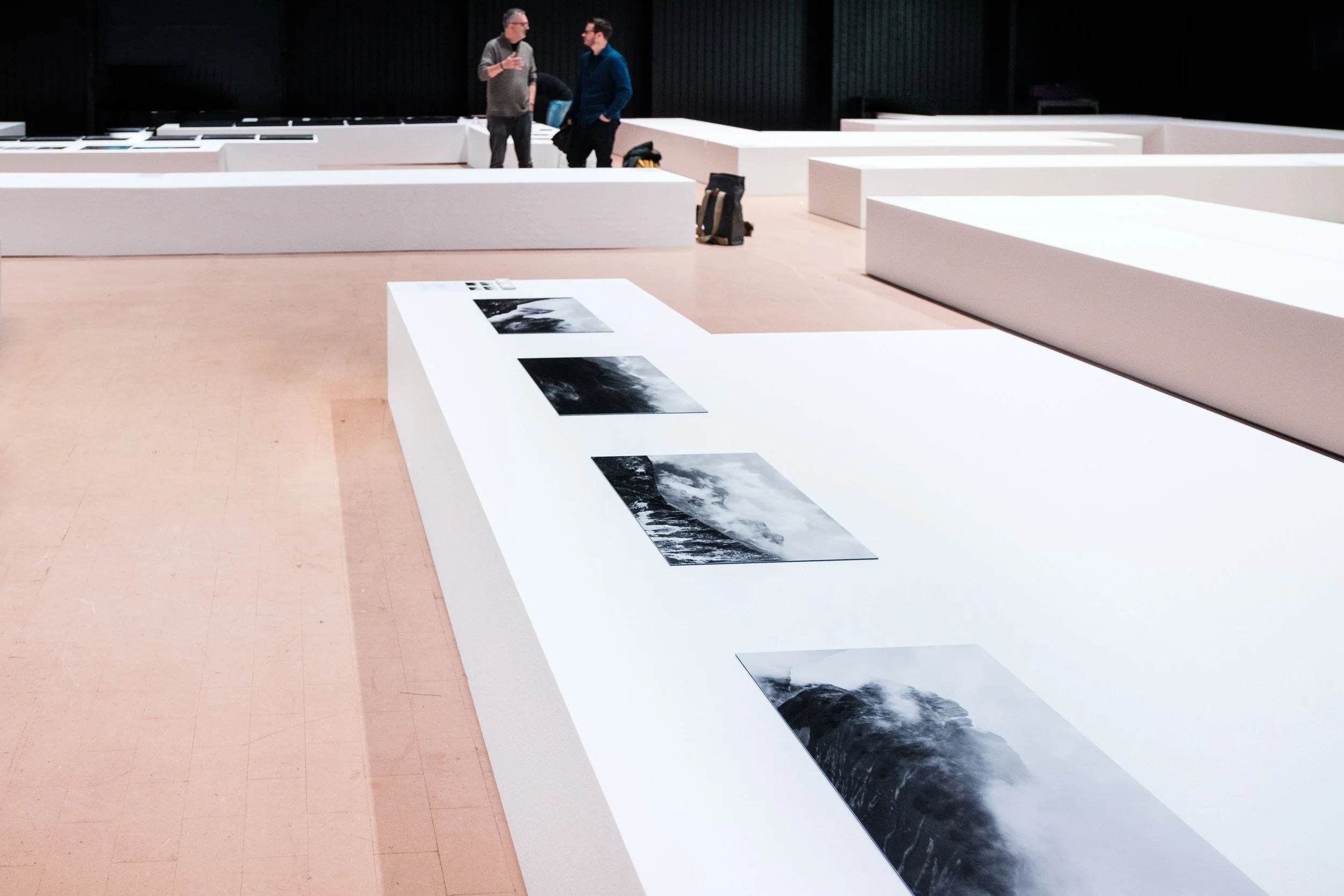 Zwei Männer sprechen miteinander in einem Ausstellungsraum mit weißen Ausstellungswänden und Schwarz-Weiß-Fotografien.
