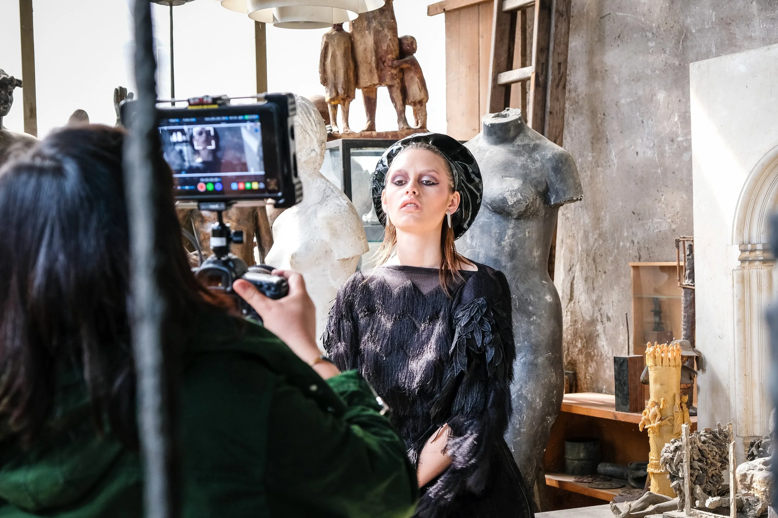 Frau posiert vor einer Kamera in einem Kunststudio, umgeben von Skulpturen und Kunstwerken.