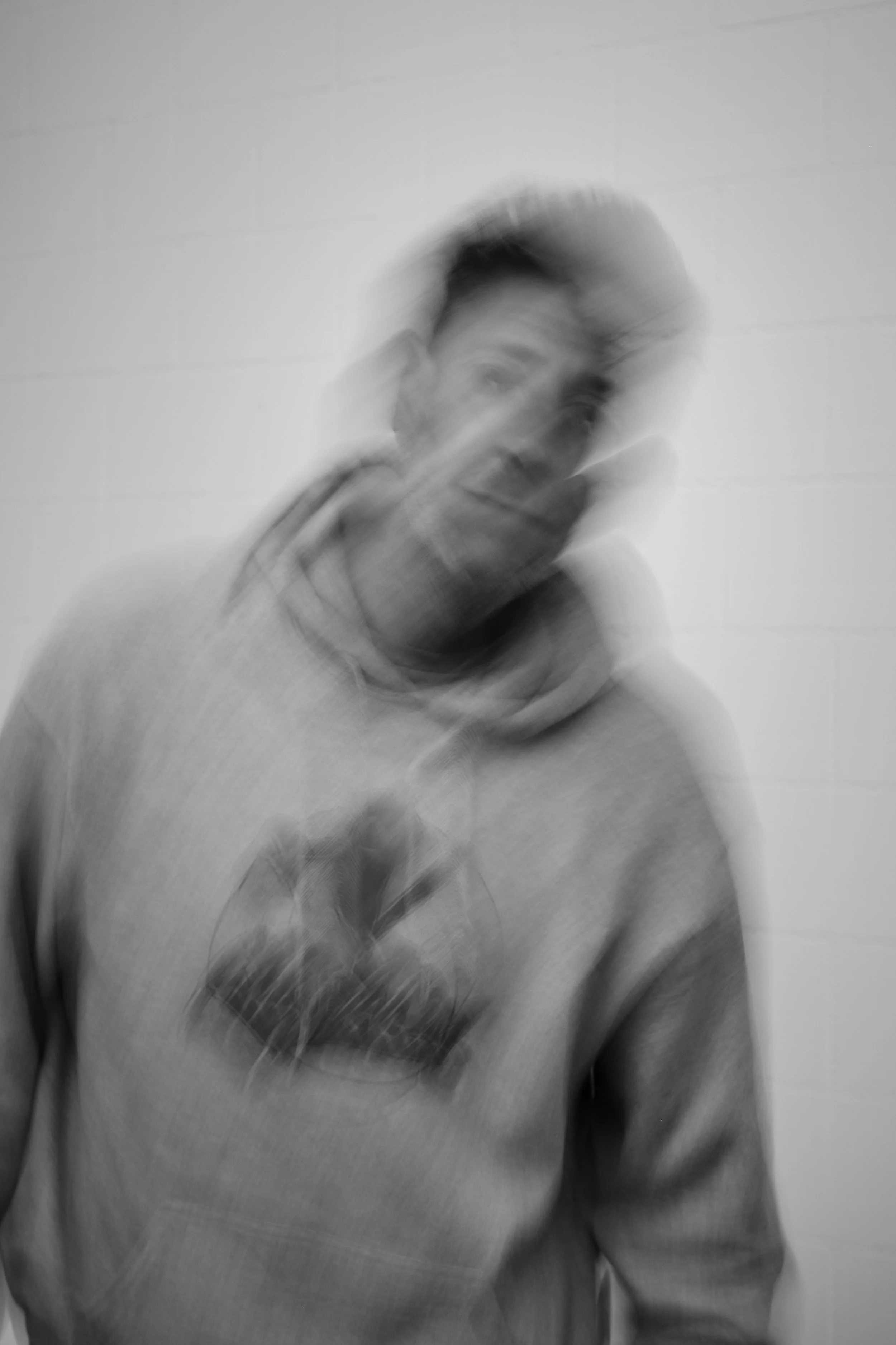 Schwarz-weiß-Bild eines Mannes mit kurzen Haaren und Hoodie, leicht verschwommen und unscharf gegen eine helle Wand.