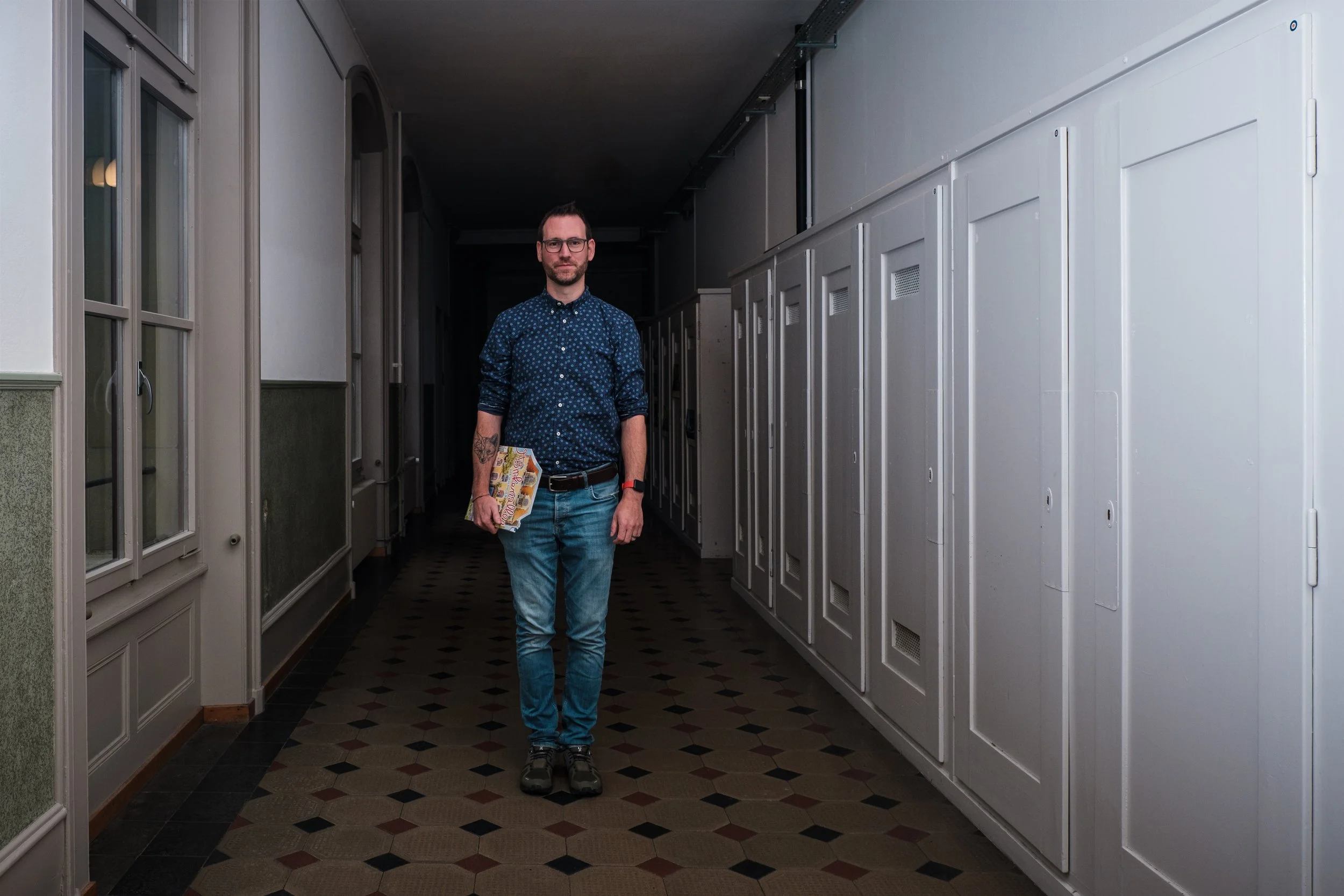 Ein Mann mit Brille, blauer gepunkteter Hemd, Jeans, steht in einem Flur mit weißen Schließfächern auf der rechten Seite und Fenstern an der linken Seite, hält eine Zeitschrift.