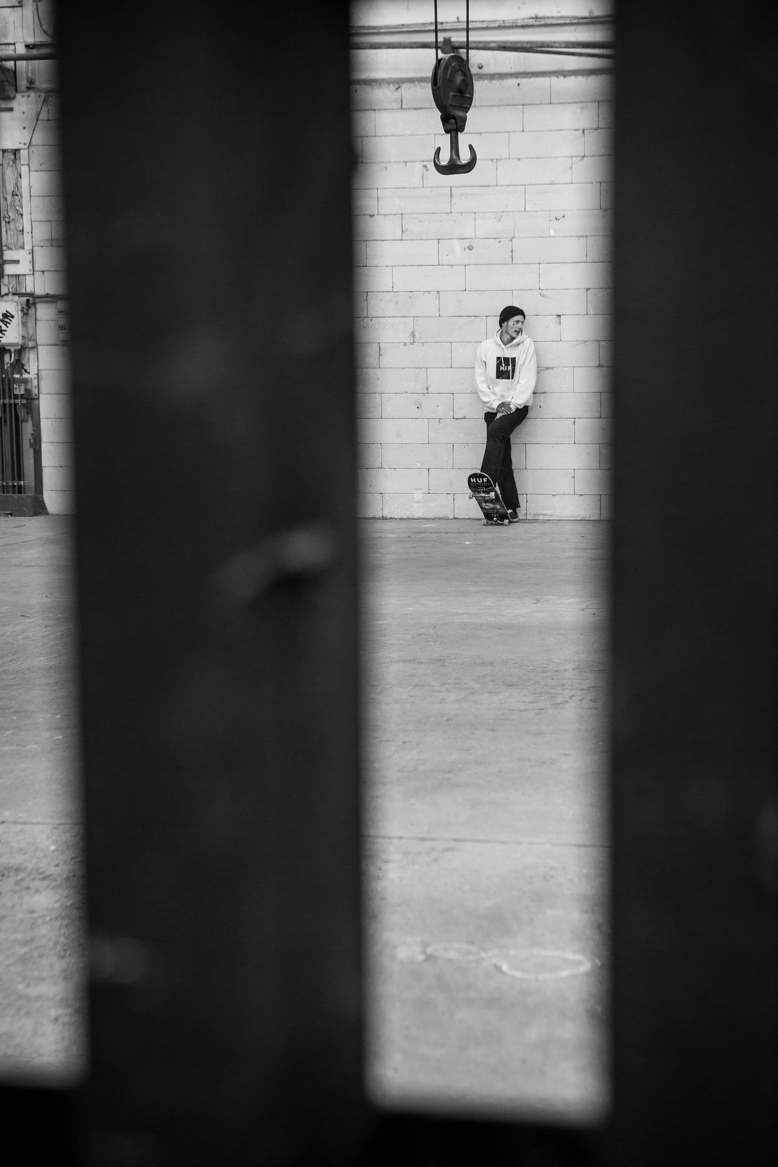 Ein Jugendlicher mit Skateboard lehnt an einer Mauer in einem indoor Skatepark, gesehen durch eine schmale Lücke zwischen dunklen Objekten.