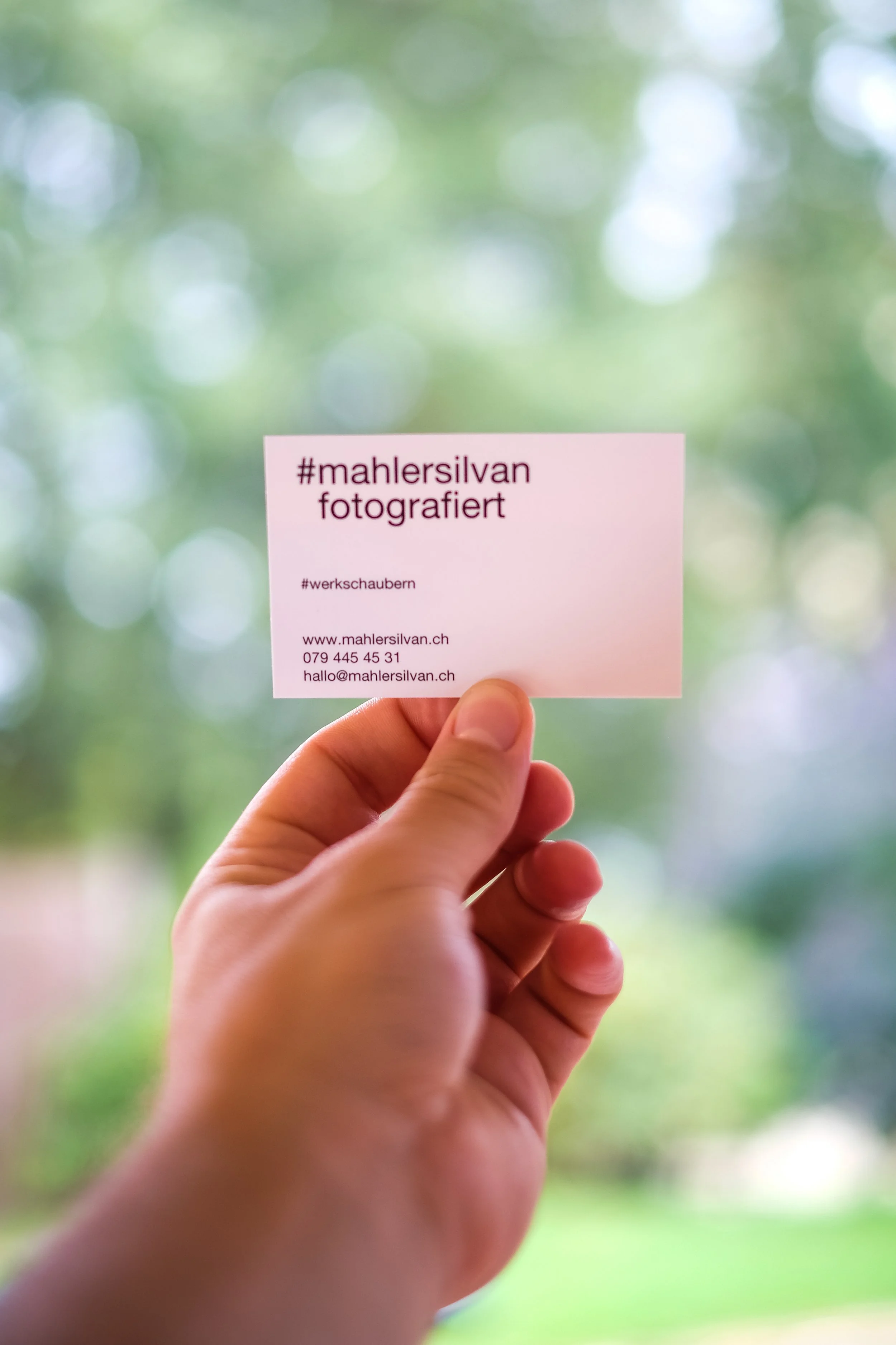 Eine Hand hält eine Kreditkarte mit dem Text '#mahlersilvan fotografiert' vor einem unscharfen, grünen Hintergrund.
