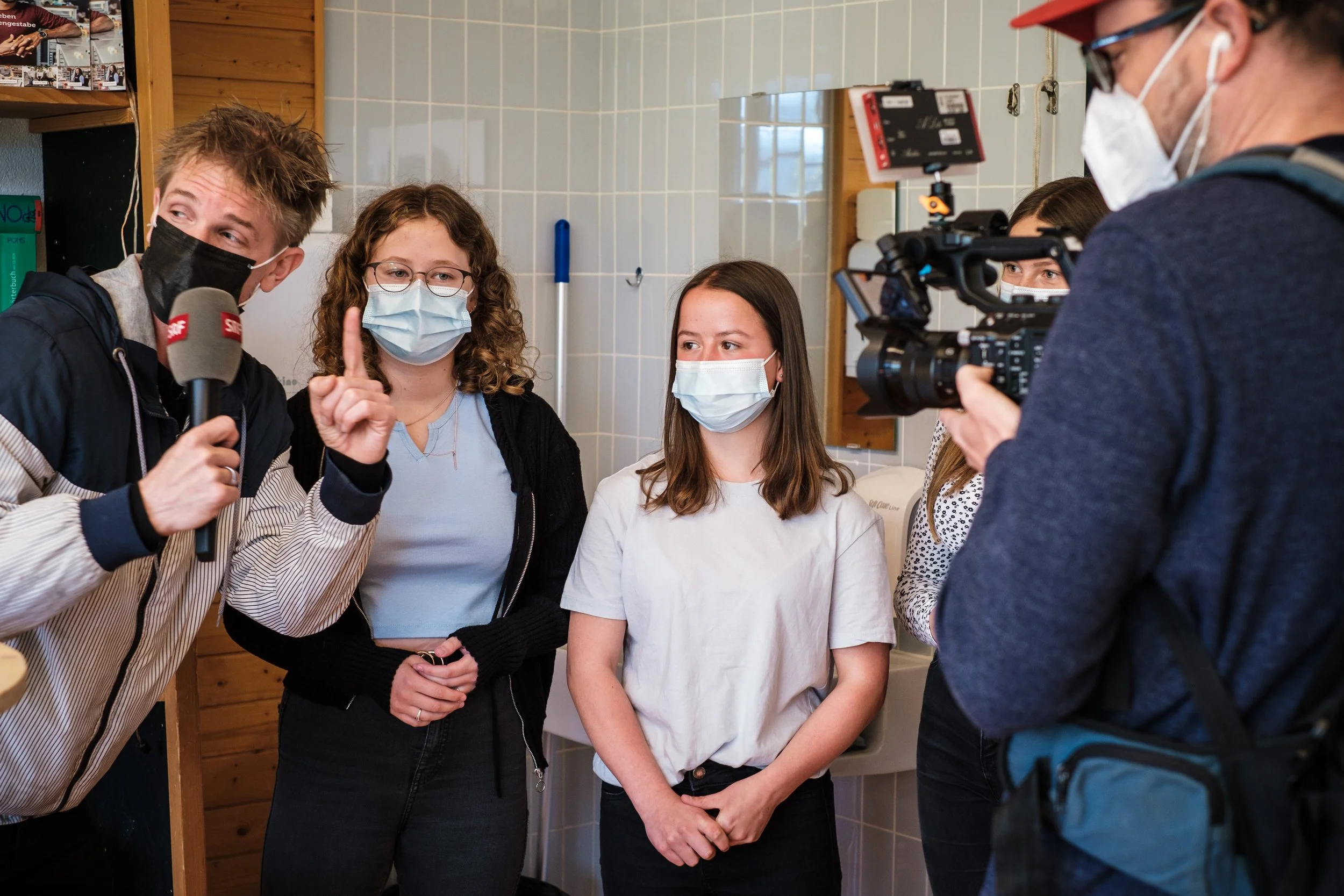 Eine Gruppe junger Menschen wird in einem Raum mit weißen Fliesen fotografiert, während ein Mann mit Kamera und Mikrofon sie interviewt. Alle tragen Gesichtsmasken.