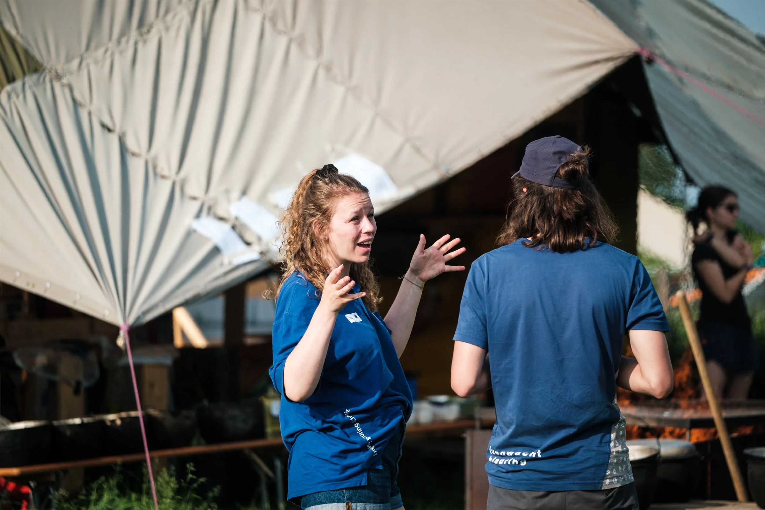 Zwei Menschen in Blue-Shirts, die im Gespräch sind, im Freien mit einem weißen Zeltdach im Hintergrund, beim Grillen oder Kochen.