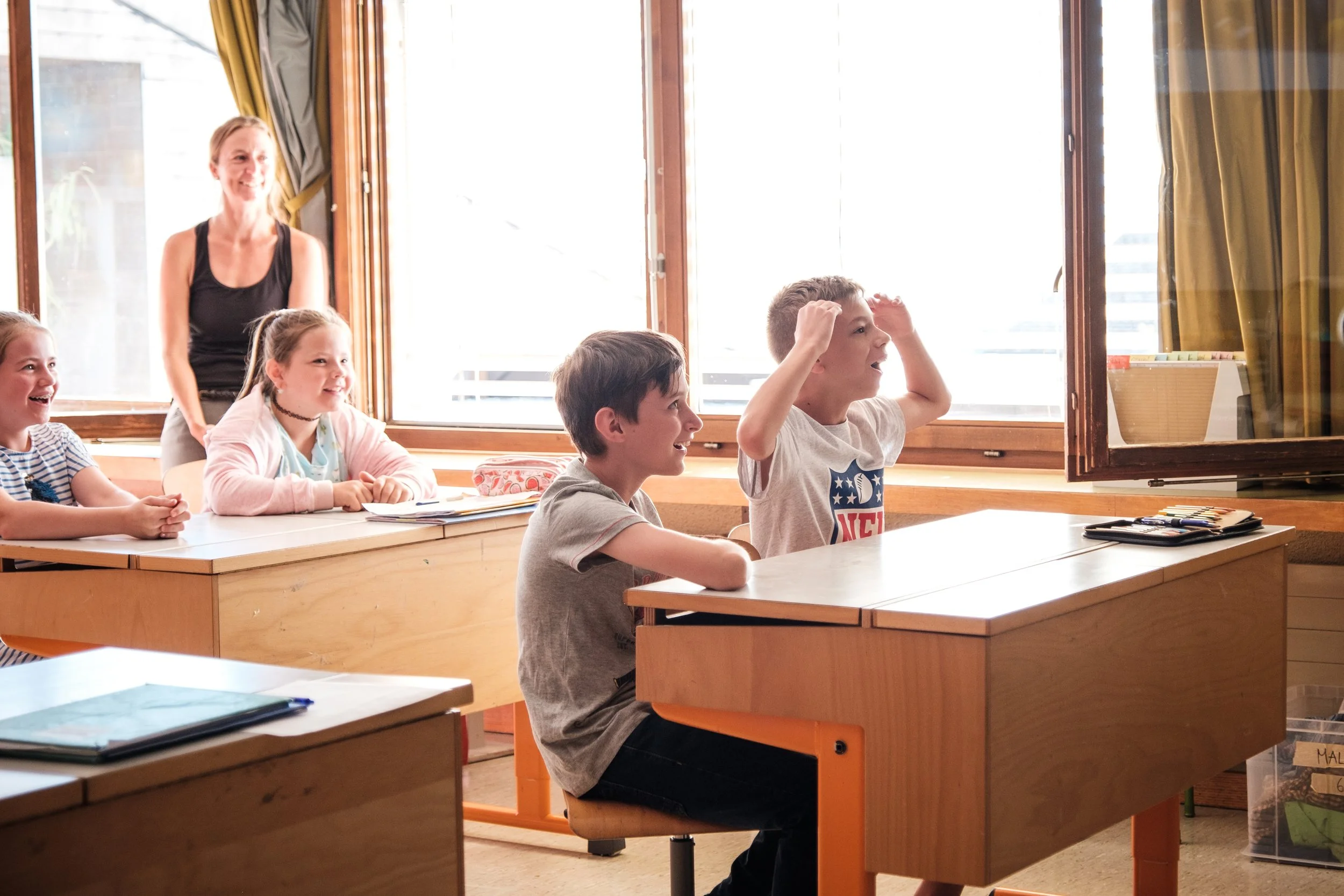 Schulklasse mit Schülern und Lehrerin, die im Klassenzimmer lachen, während ein Junge seinen Kopf mit den Händen hält.