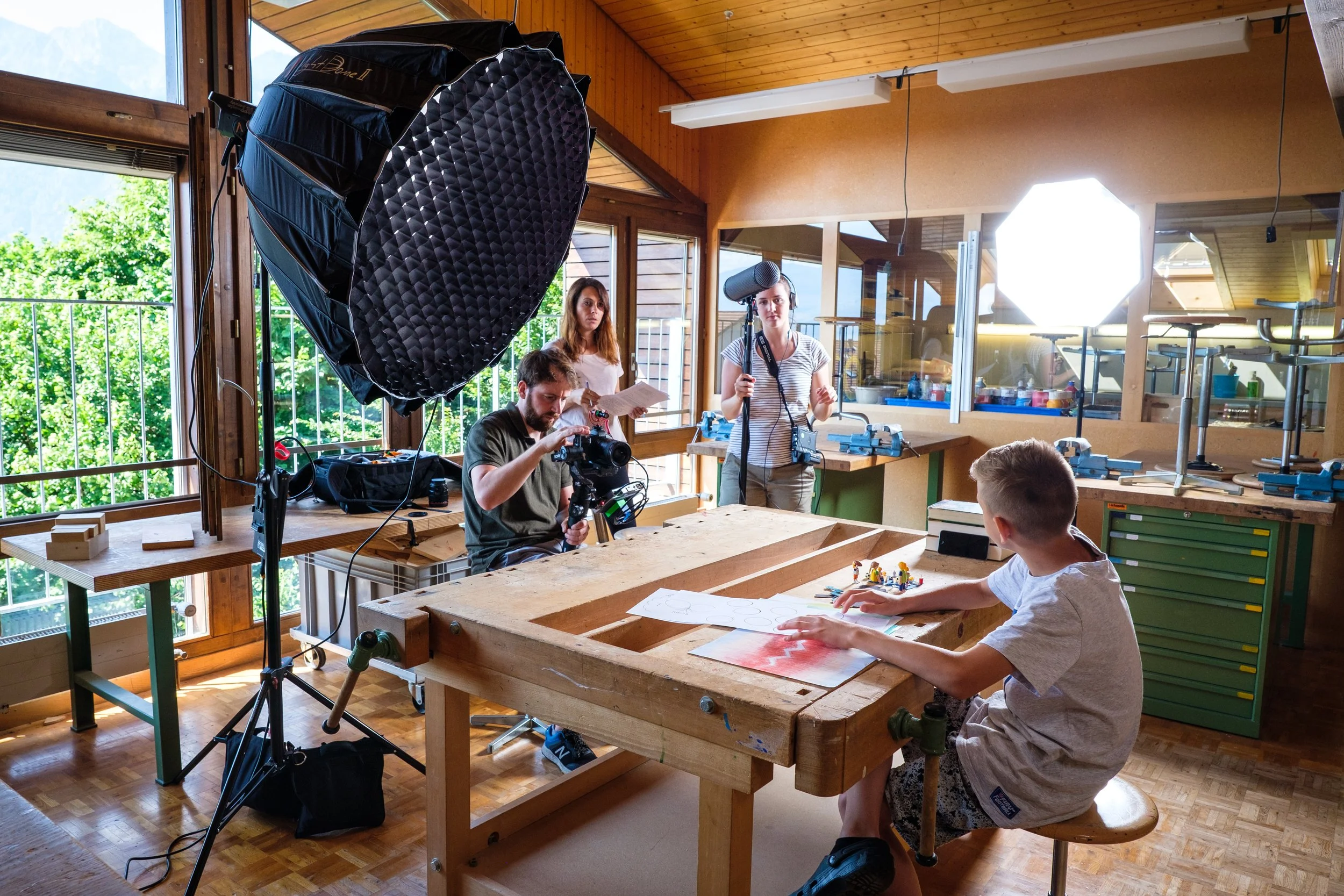 Ein Filmteam macht in einem hellen, mit Holz verkleideten Raum Filmaufnahmen eines Jungen, der an einem Arbeitstisch mit Lego Figuren spielt. Es gibt ein großes, professionelles Licht, eine Kamera, eine Person mit einem Mikrofon und eine Person mit e