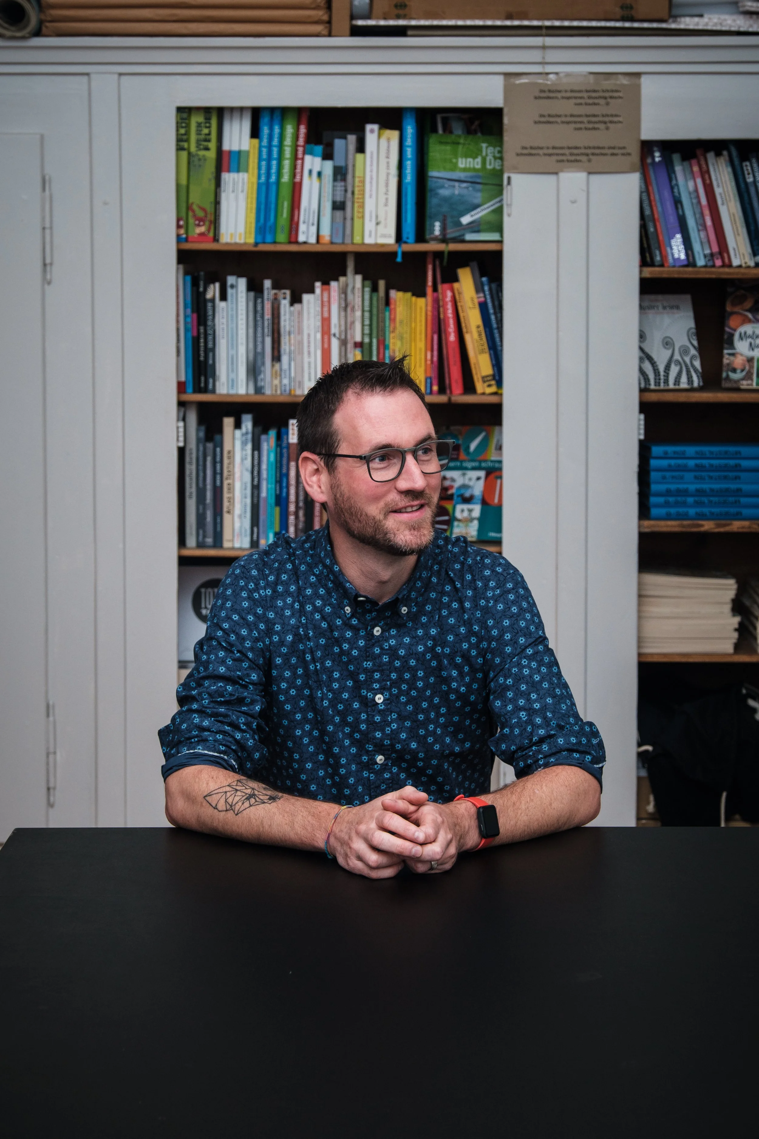 Ein Mann mit Brille und Bart sitzt an einem Tisch vor einem Bücherregal.