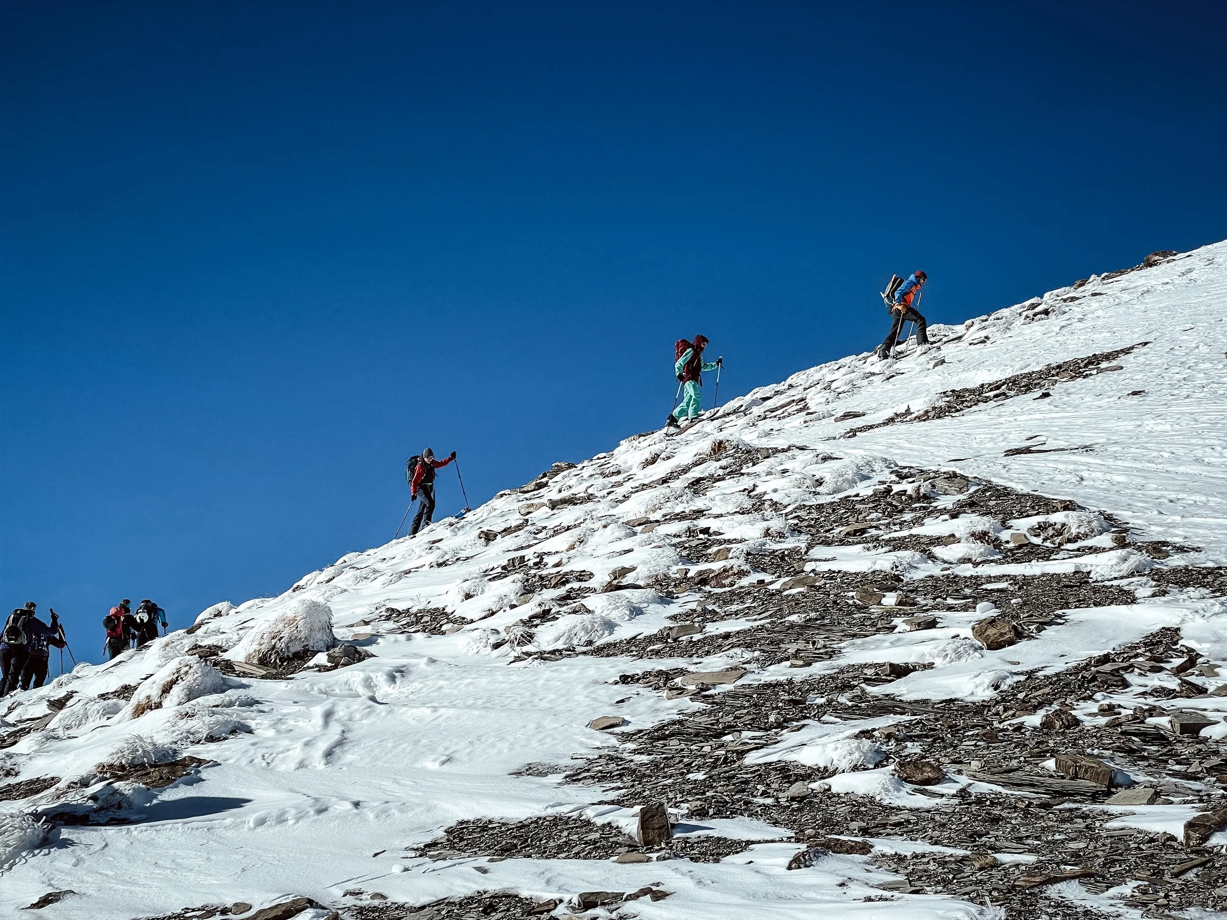 Menschen steigen in einer Schneelandschaft bergauf, einige mit Rucksäcken und Wanderstöcken, unter klarem blauen Himmel.