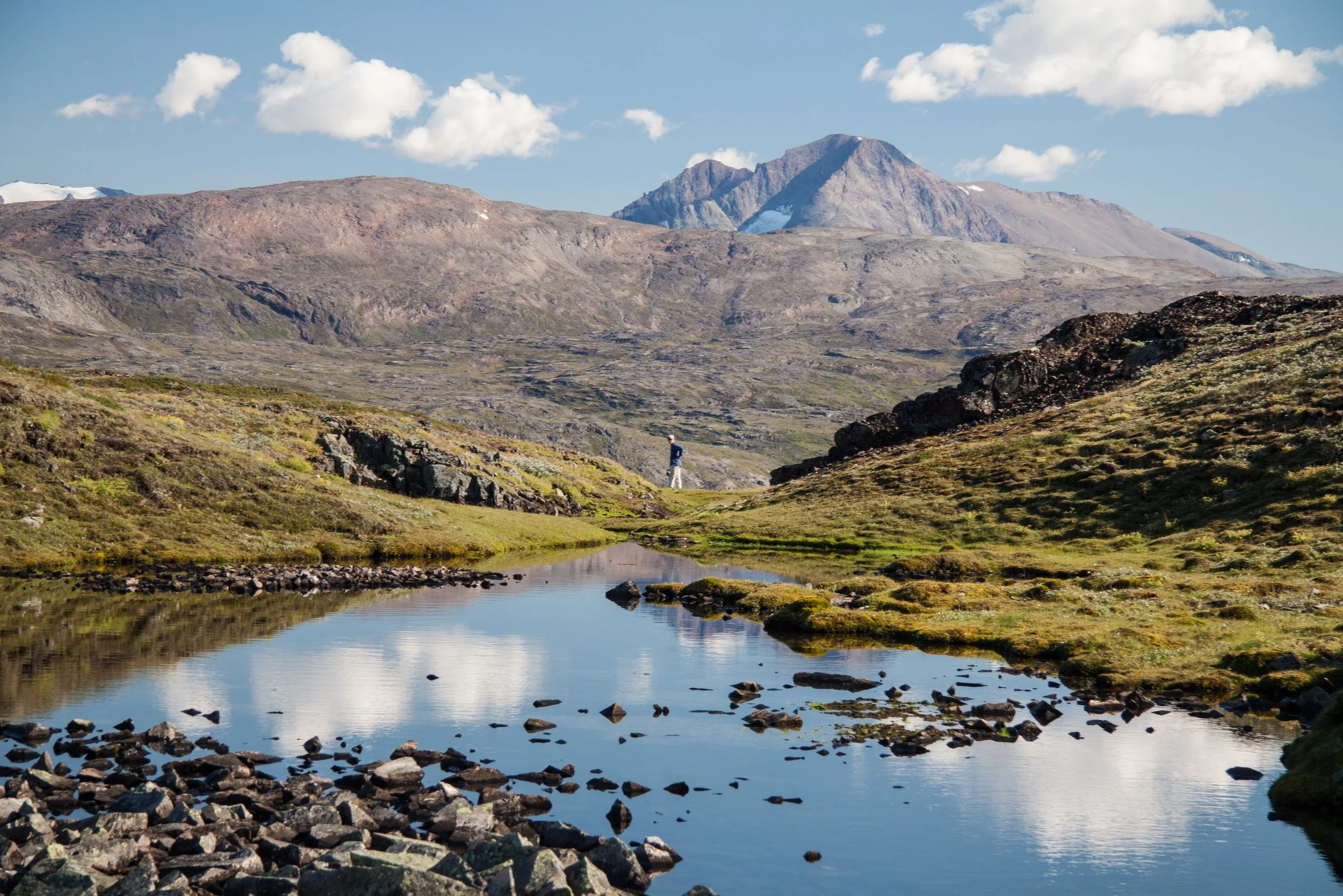Berglandschaft mit schimmerndem Fluss, felsigen Hügeln und hohen Bergen im Hintergrund, blauer Himmel mit wenigen Wolken.
