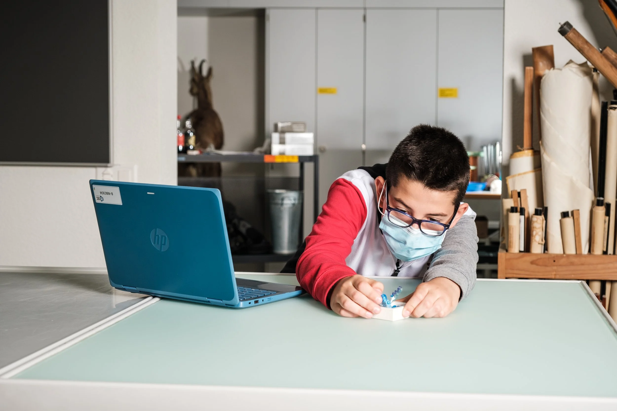 Ein Junge mit Brille und Maske arbeitet an einem Projekt in einem Raum, auf dem Tisch liegt ein Laptop und kleine Objekte. Im Hintergrund sind Regale und ein Geweih an der Wand.