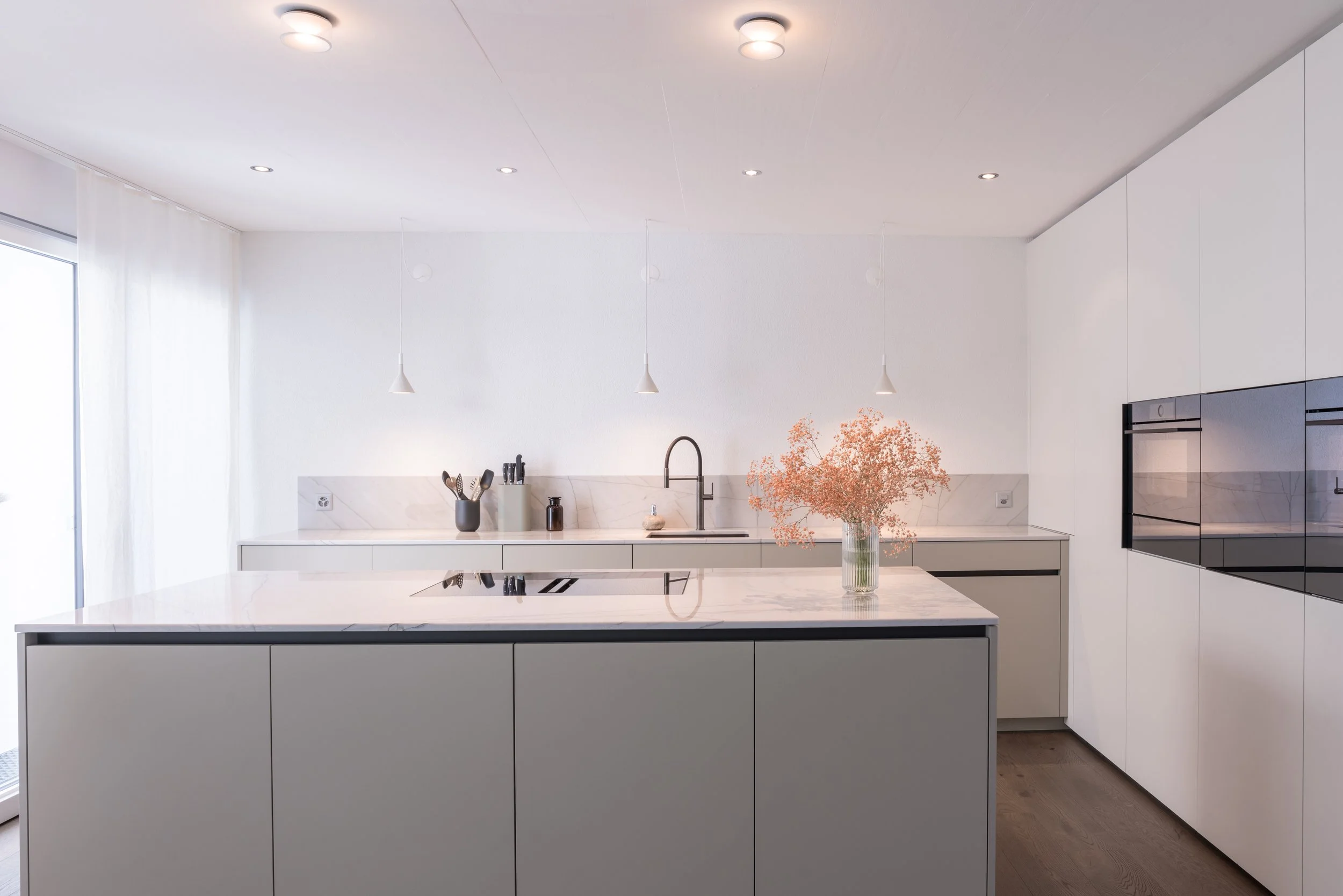 Moderne, minimalistische weiße Küche mit atemberaubender Sicht auf den Balkon, Kücheninsel mit weißen Schränken und Marmor-Arbeitsplatte, Vase mit Trockenblumen, Backofen in der Wand eingelassen, und kleine Küchenutensilien auf der Arbeitsfläche.