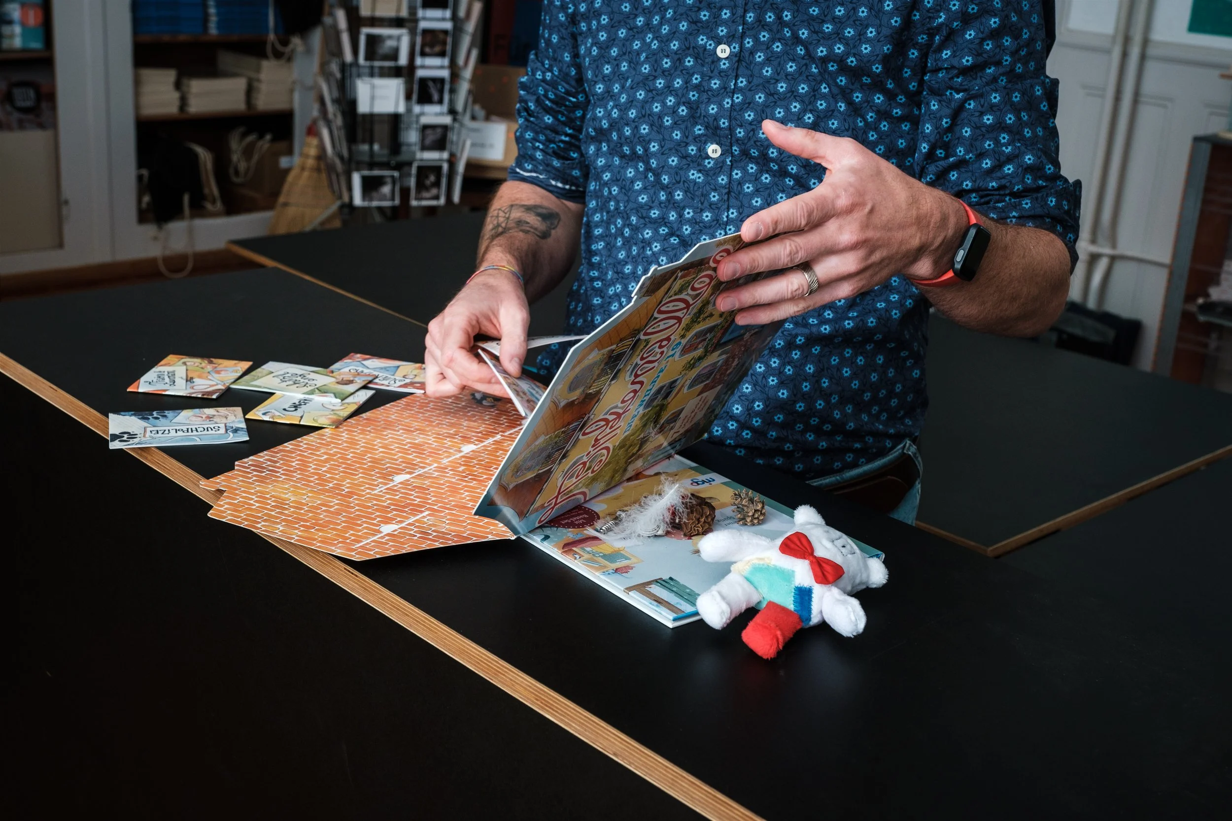 Person betrachtet ein aufgeschlagenes Scrapbook mit Fotos, Bastelmaterial und einem kleinen Teddy an einem Tisch in einem Raum mit Bücherregalen.