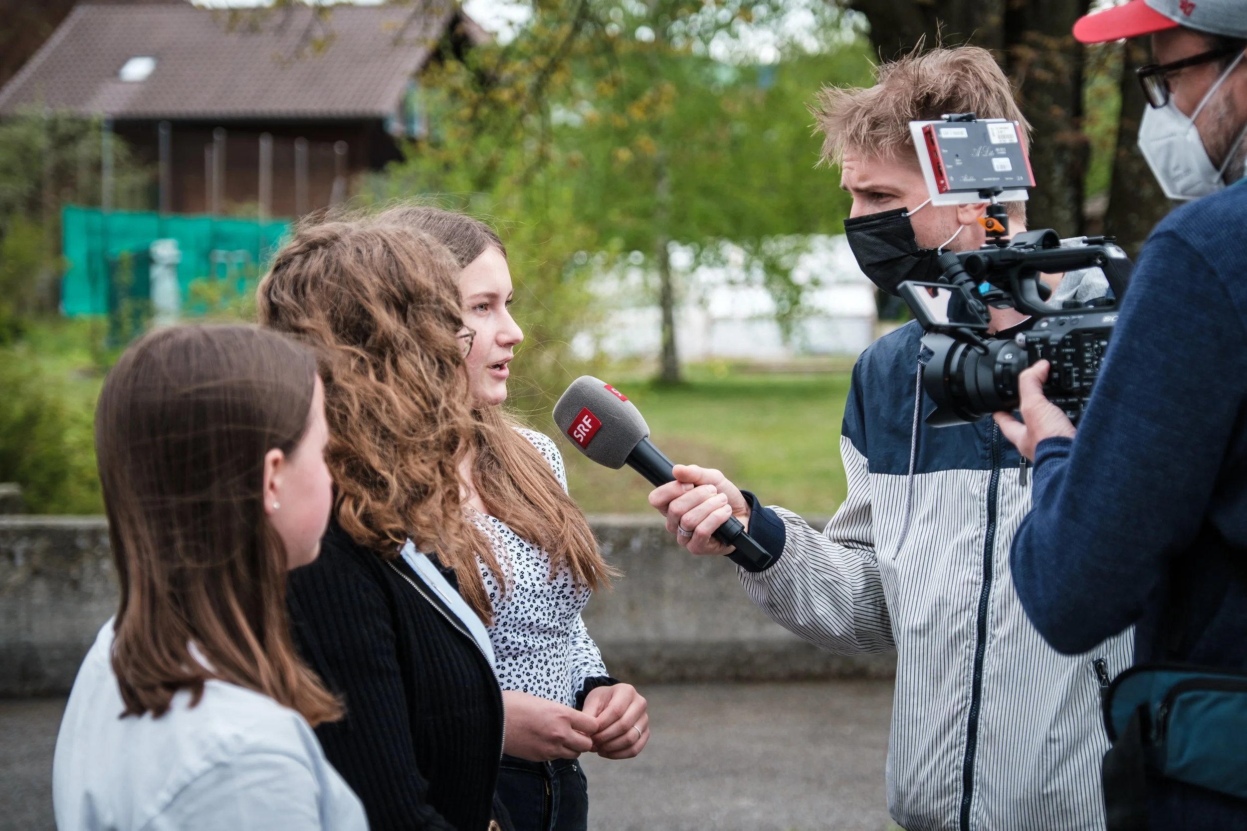Drei junge Frauen im Gespräch mit einem Reporter, der eine Kamera und ein Mikrofon mit SRF-Logo hält, während ein Kameramann filmt. Alle tragen Mundmasken.