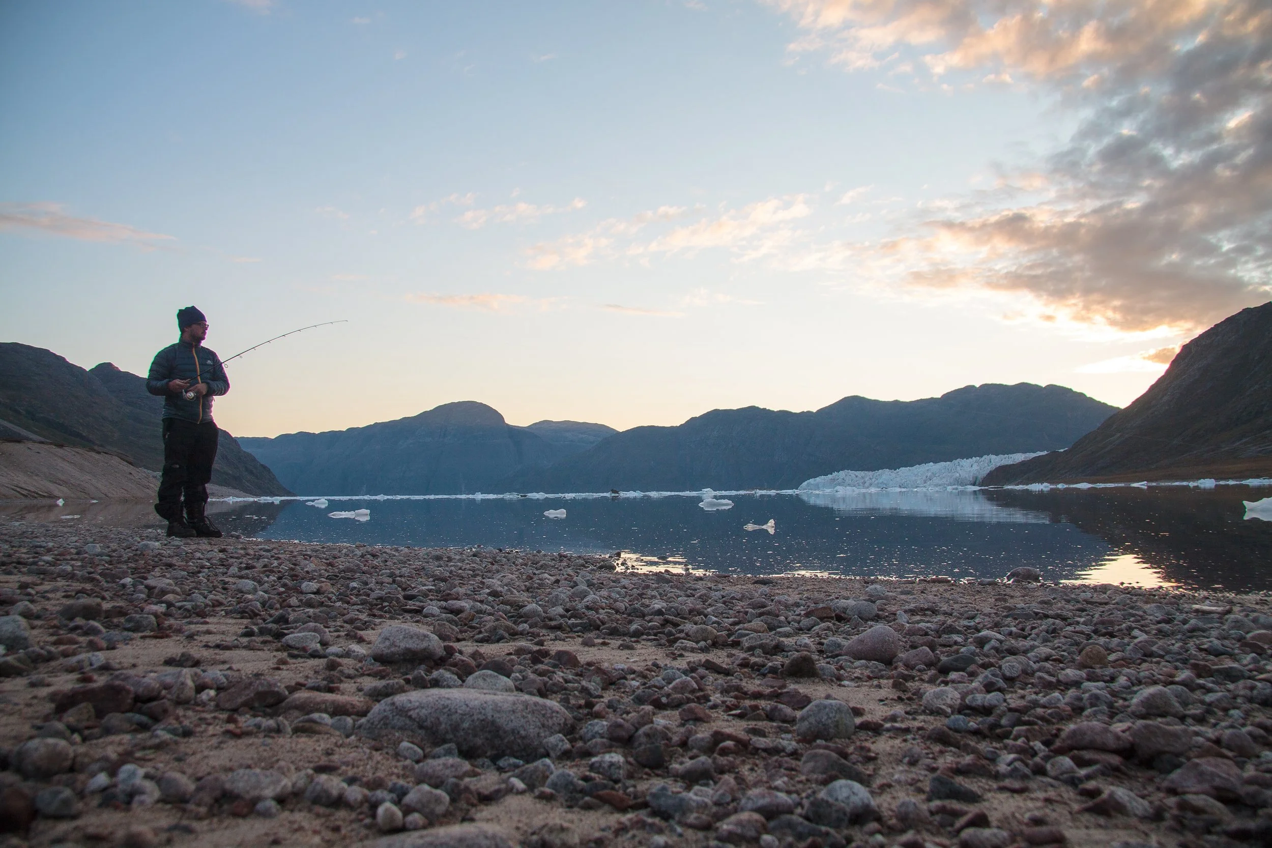 Ein einzelner Angler steht am Ufer eines Fjords mit Bergen im Hintergrund. Es ist Dämmerung, der Himmel ist teils bewölkt, und Eisstücke schwimmen im Wasser.