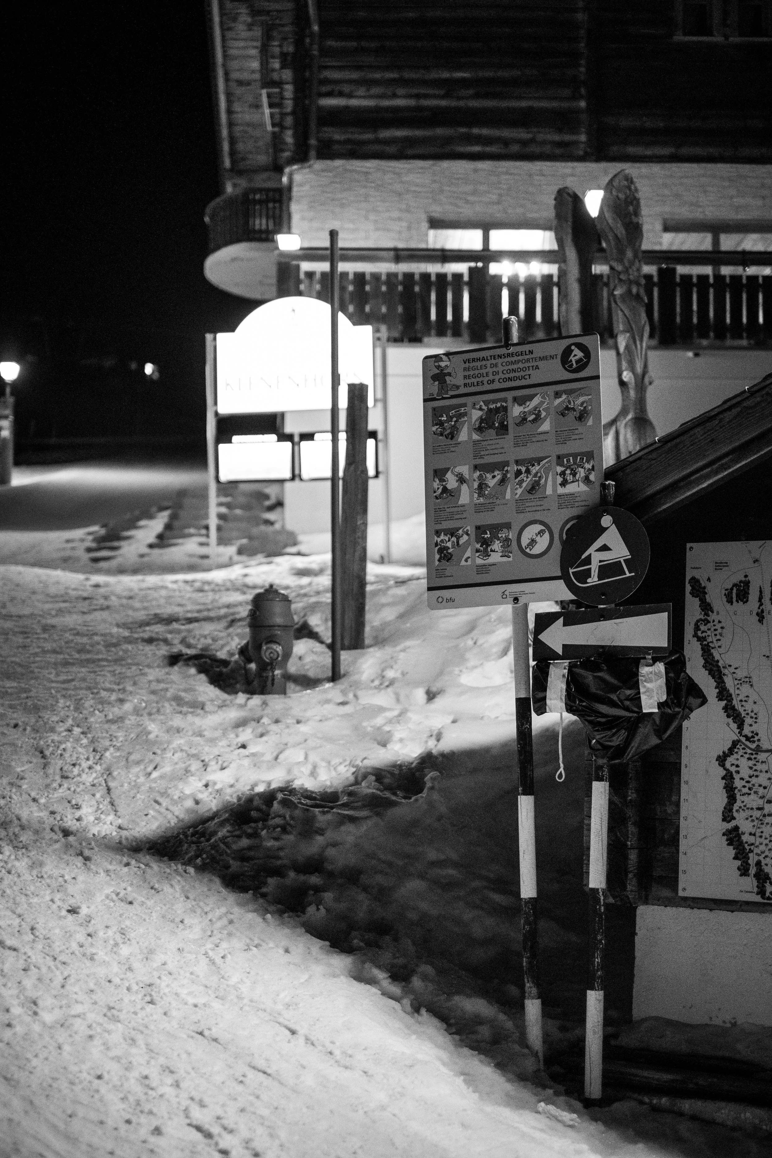 Schneebedeckte Straße bei Nacht mit Beschilderung, einem Feuerhydranten und einer Holzhütte im Hintergrund.