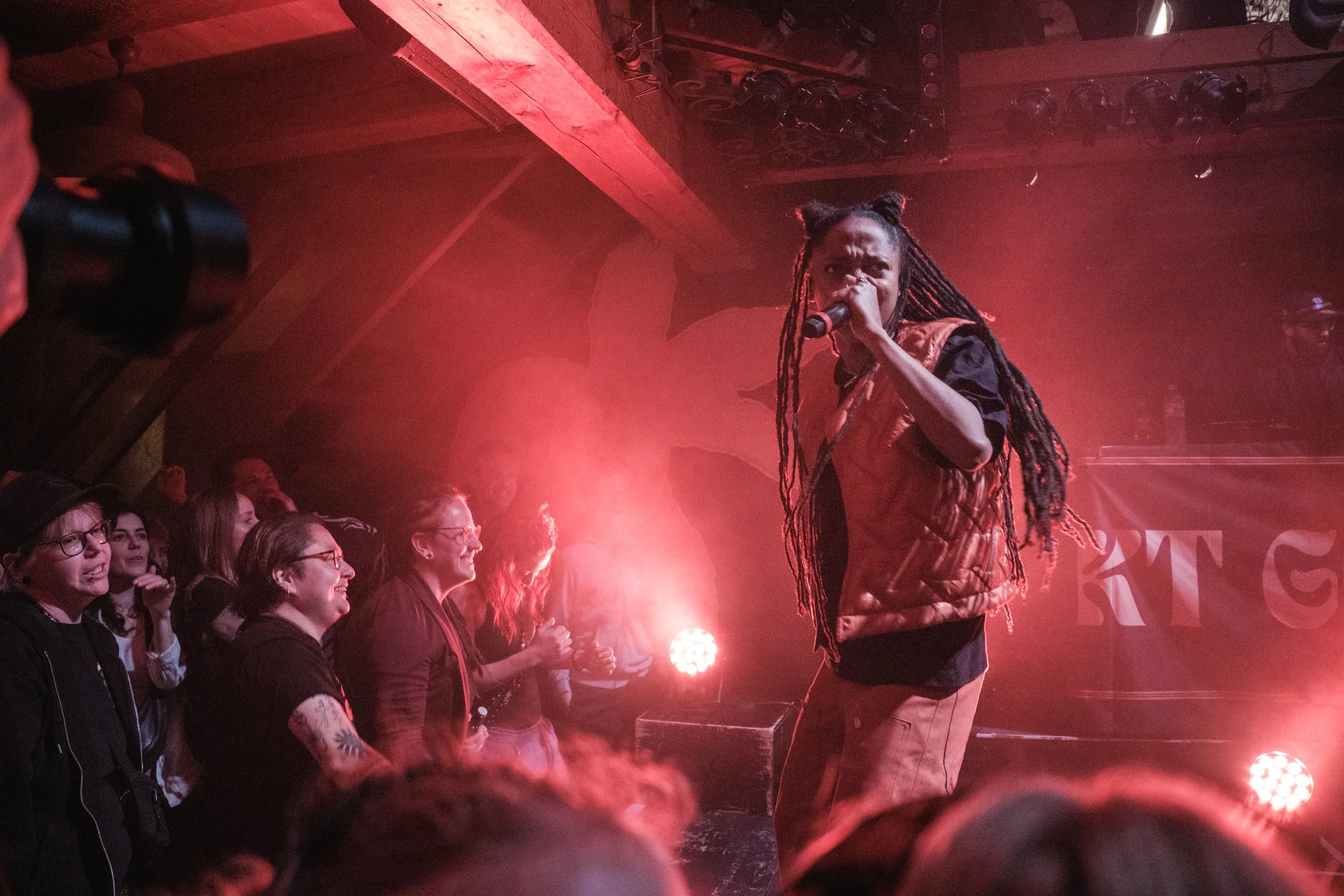 Eine Musikerin singt in einem Club vor einem Publikum, das begeistert zuschaut. Sie trägt einen braunen Weste und hat lange Zöpfe. Es ist dunkel, und es gibt rote Bühnenlichter.