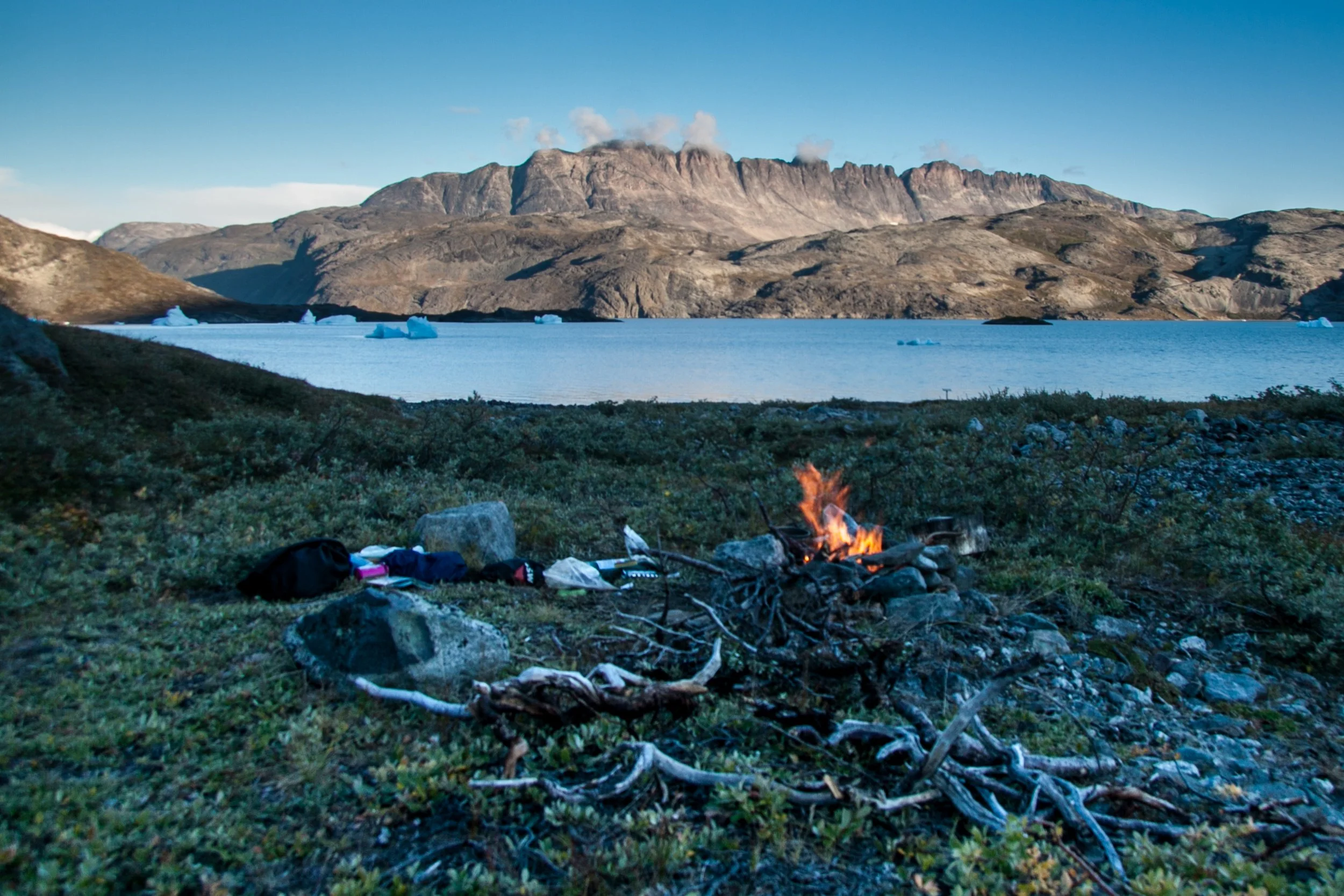 Lagerfeuer auf einer Wiese vor einem See, umgeben von Bergen mit Gletschern, bei Dämmerung