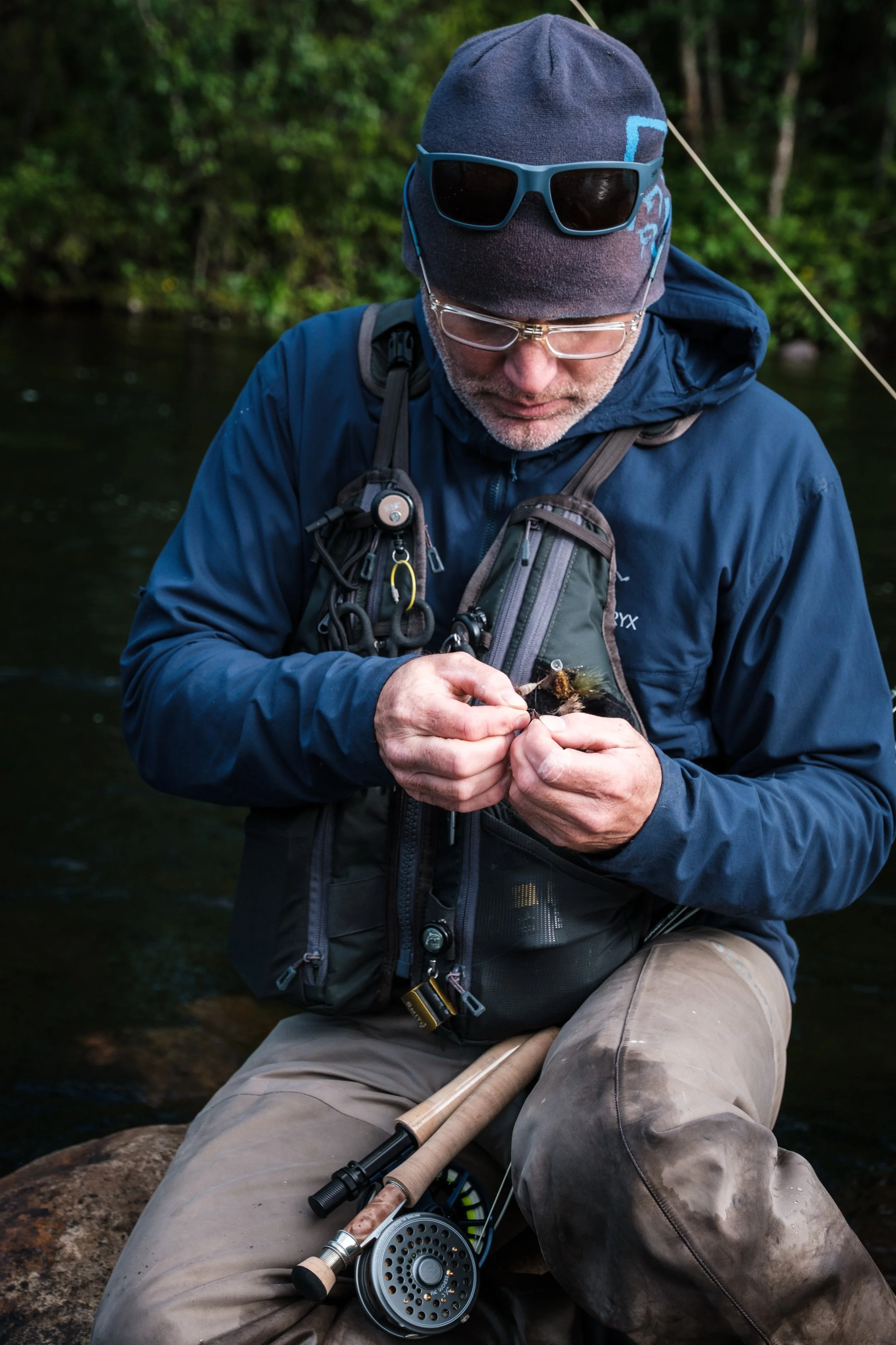 Ein Mann beim Fliegenfischen im Fluss, trägt eine Mütze, Brille und eine Jacke, sitzt auf einem Stein und bereitet einen Fischerköder vor.