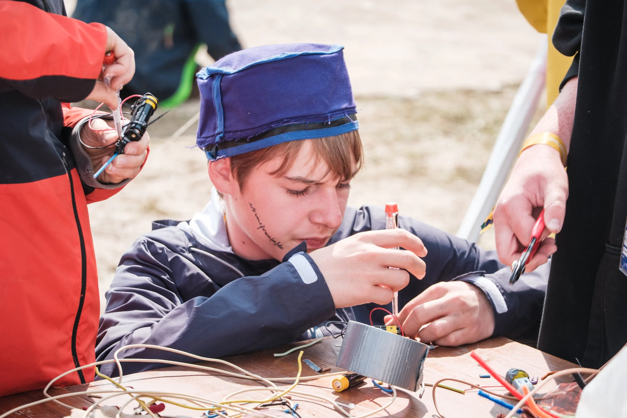 Ein junger Mensch arbeitet mit elektronischen Bauteilen und Drähten auf einem Tisch, während er von mehreren Personen unterstützt wird.