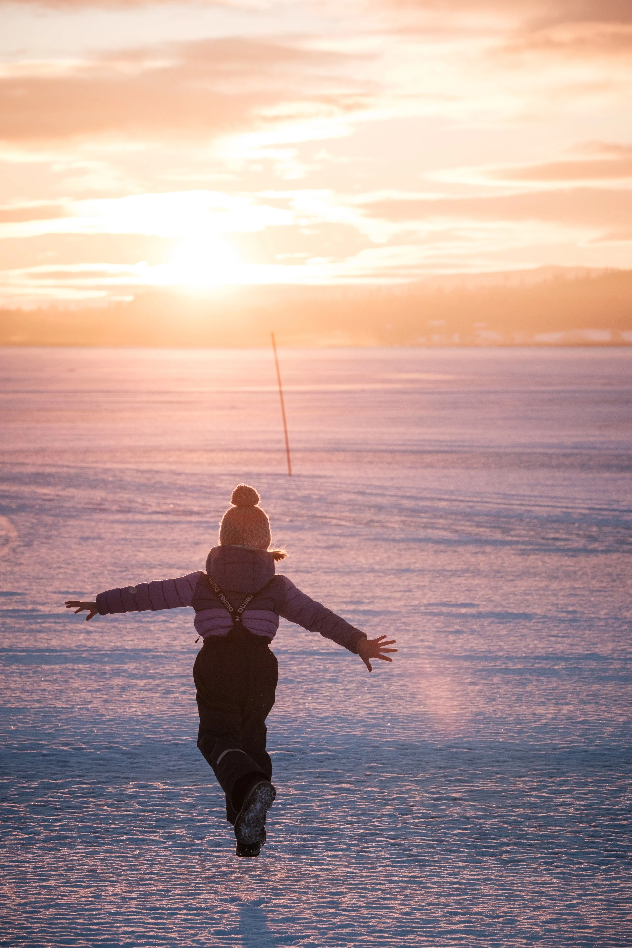 Ein Kind, das im Schnee läuft, trägt eine Winterjacke, Mütze und Handschuhe, mit Sonnenuntergang im Hintergrund.