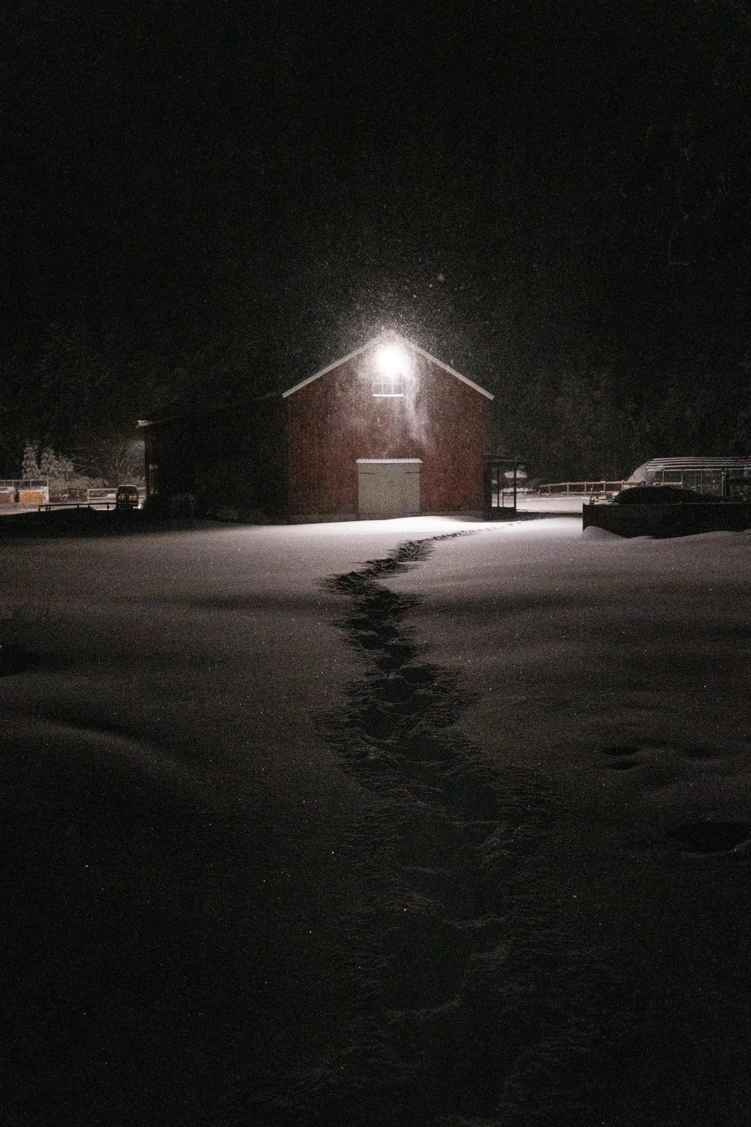 Schneebedecktes Land bei Nacht mit einem roten Stallgebäude, das von einem hellen Licht beleuchtet wird, und Fußspuren im Schnee