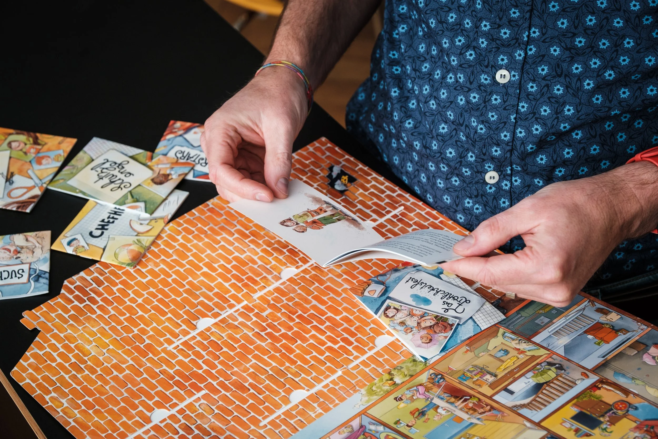 Eine Person spielt ein Brettspiel mit verschiedenen Komponenten, darunter Karten, ein Spielbrett in Ziegelsteinoptik und andere Spielmaterialien auf einem schwarzen Tisch.