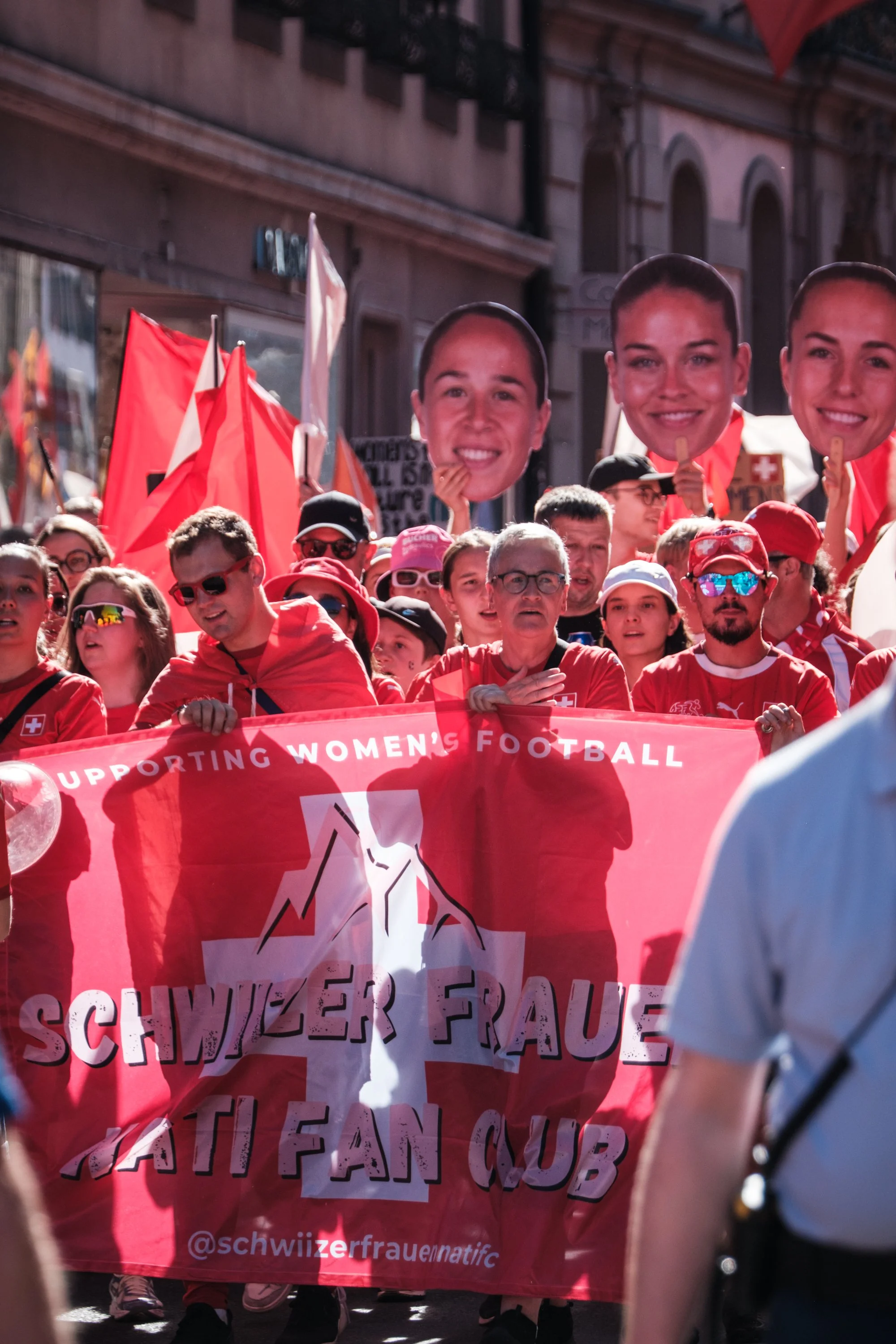 Menschen nehmen an einer Demonstration für Frauenfußball in der Schweiz teil, halten Schilder und Banner, tragen rote Kleidung, inmitten einer Menschenmenge auf einer Stadtstraße bei Tageslicht.