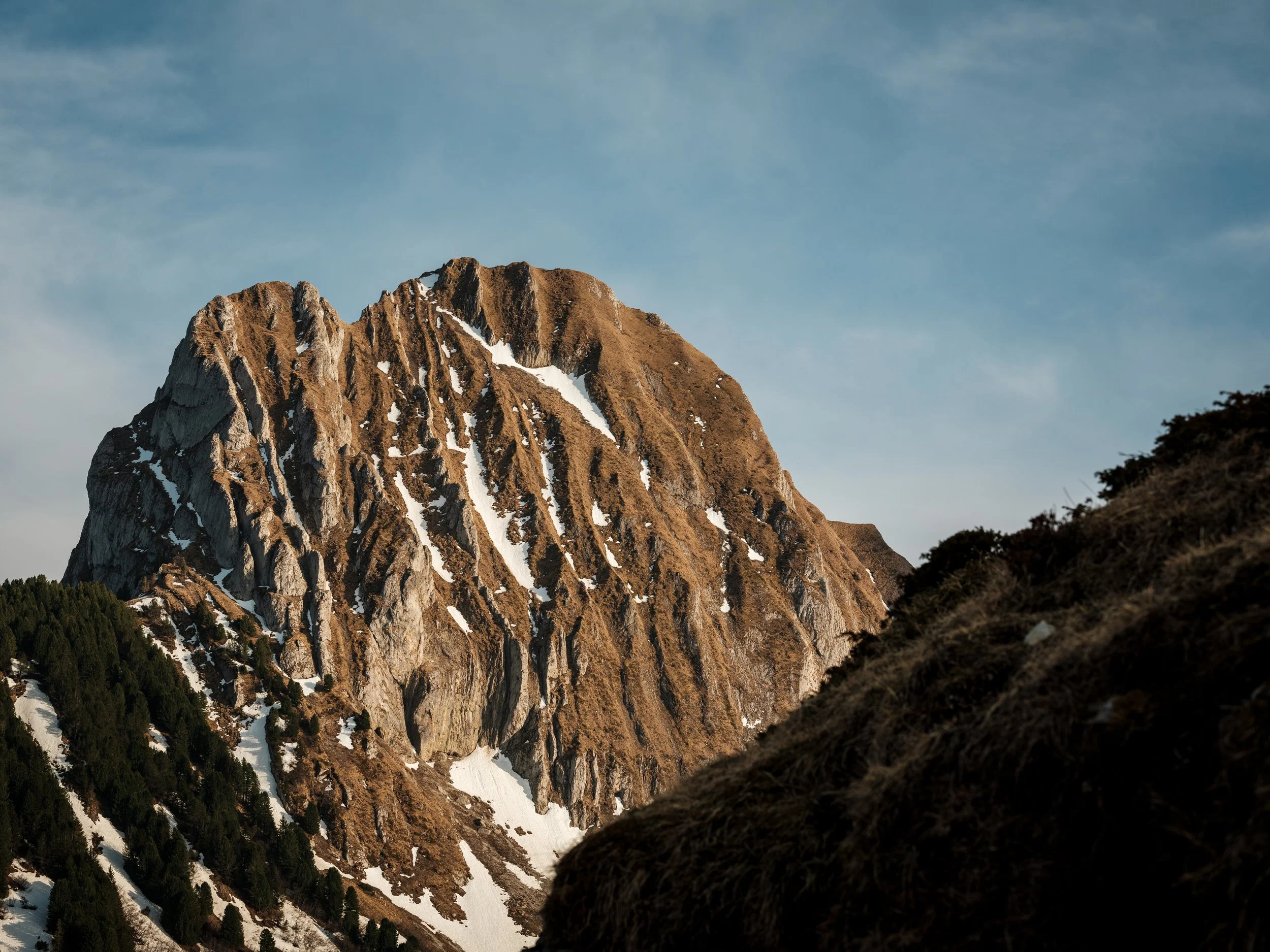 Berg mit Felsen und Schneespuren bei Sonnenuntergang, bewölkter Himmel im Hintergrund.