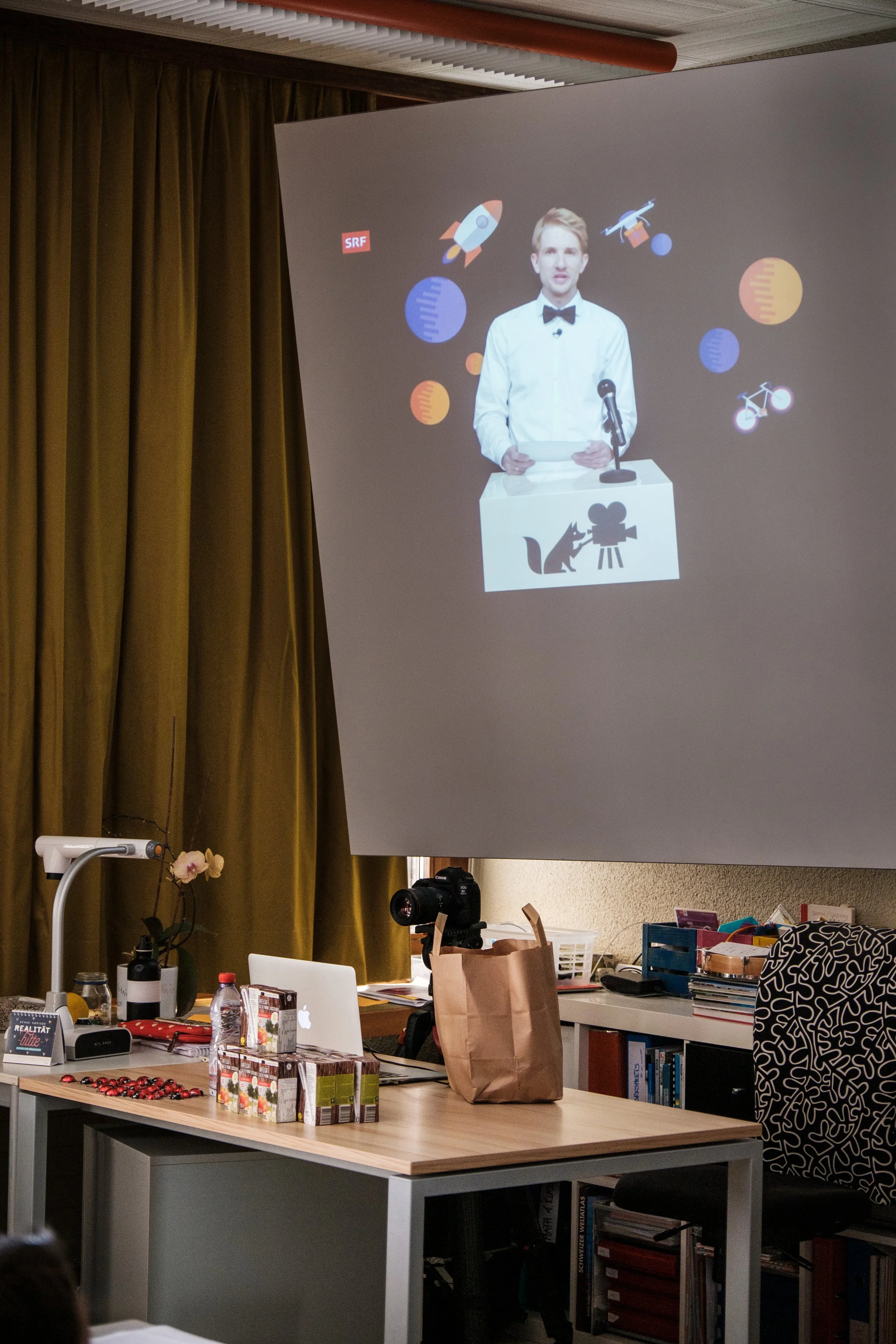 Ein Mann mit Smoking und Fliege spricht bei einer Präsentation auf einer Leinwand, die Raumfahrt- und Wissenschaftssymbole zeigt.