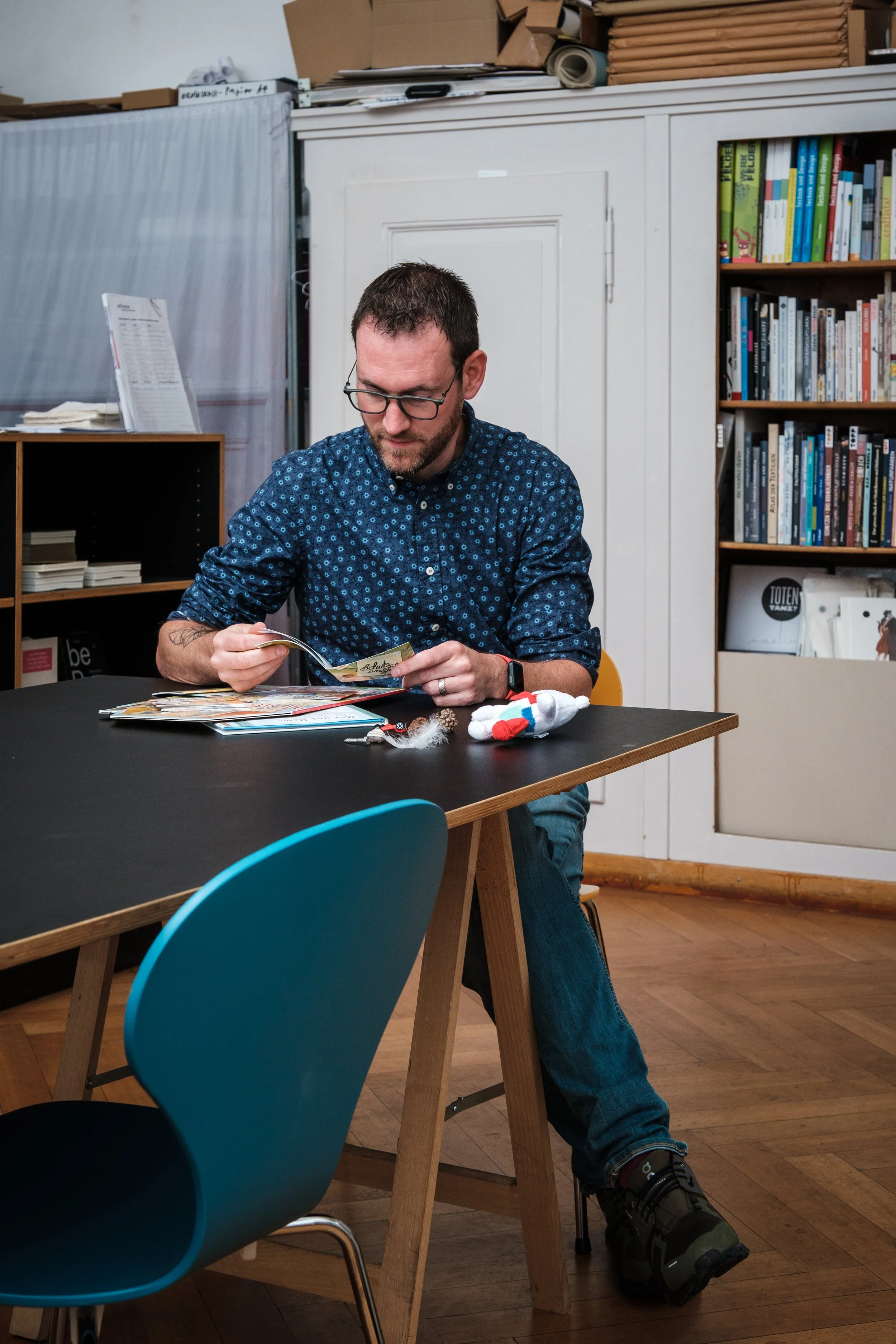 Ein Mann sitzt an einem Tisch in einem Raum mit Bücherregalen, liest Papiere und hat verschiedene Gegenstände vor sich, darunter ein Kuscheltier. Er trägt eine Brille, ein blauen Hemd mit Muster und eine Uhr.