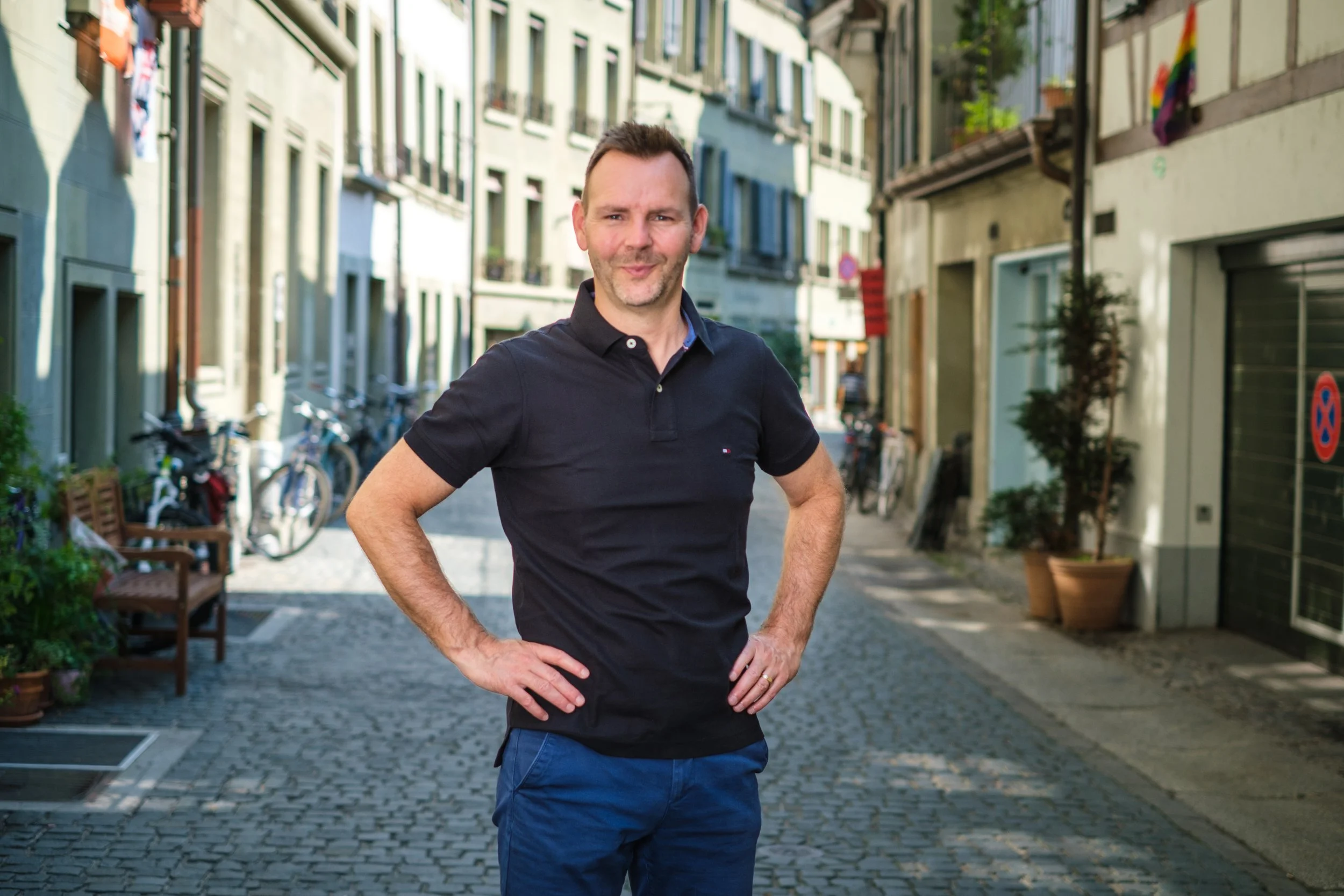 Ein Mann steht auf einer Kopfsteinpflasterstraße, umgeben von mehrstöckigen Gebäuden, Fahrrädern und Pflanzen, mit einer Hand an der Hüfte, lächelnd in die Kamera schauend.