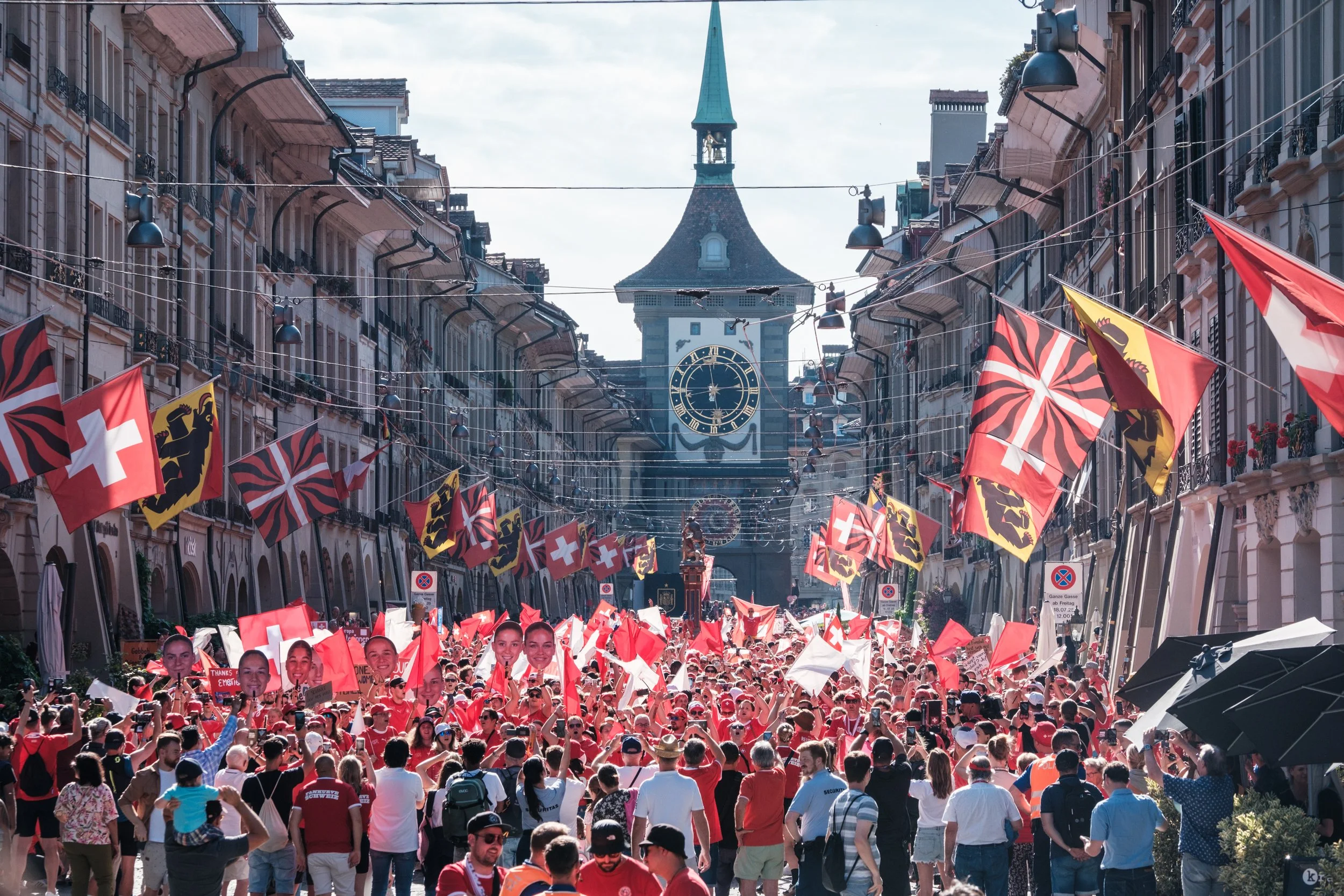 Menschen versammeln sich bei einer Straßenveranstaltung in der Schweiz mit Schweizer- und Kantonalfahnen, vor einer großen Uhr auf einem Turm.